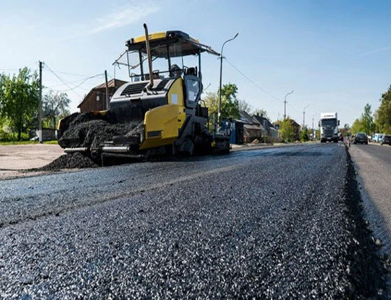 Asphalt paving machine laying fresh black asphalt on a road in a rural area with houses and vehicles visible in the background.