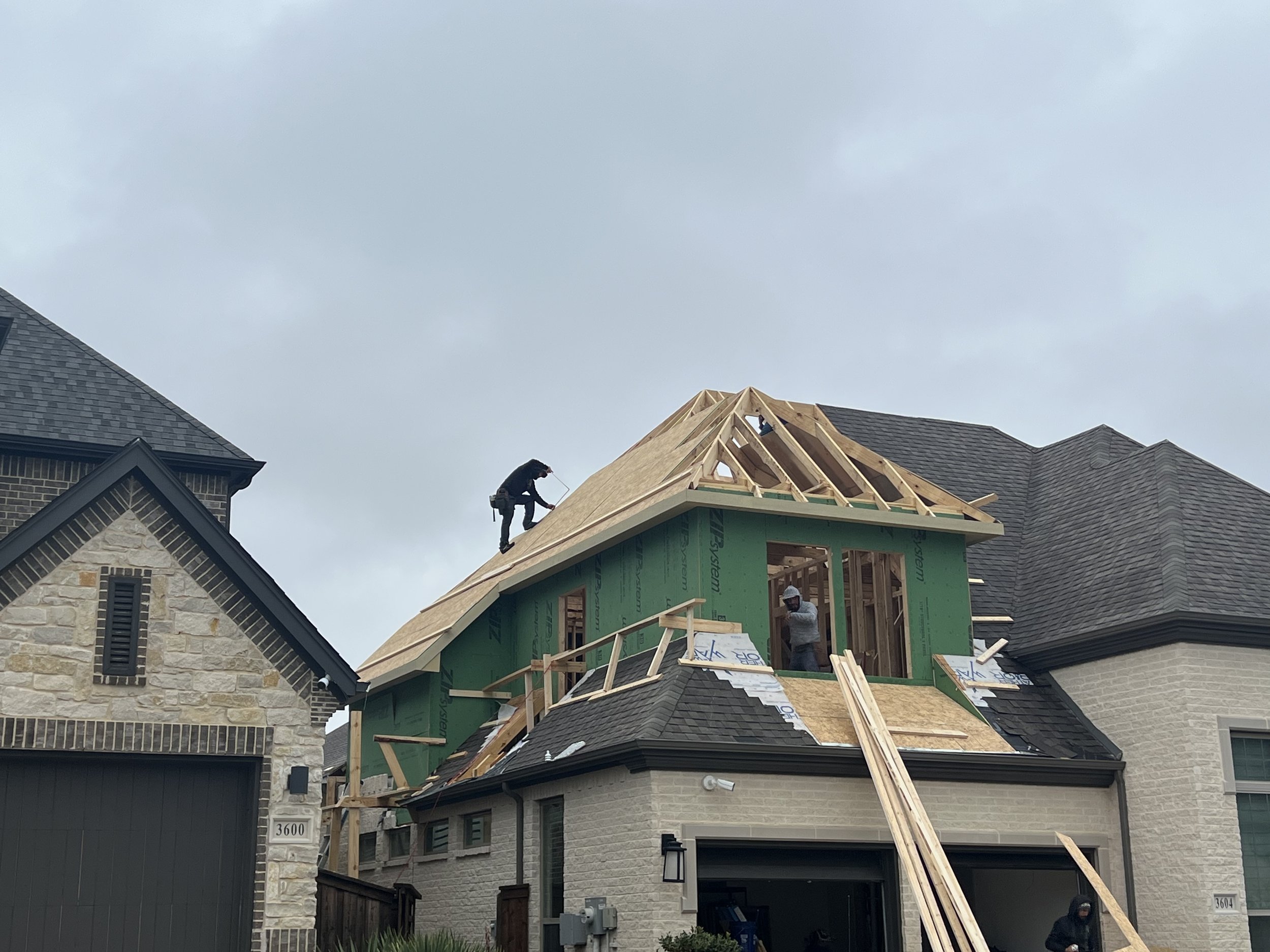 Construction workers building a second story on a house, with one worker working on the roof and two inside the structure, surrounded by wooden framing and planks.