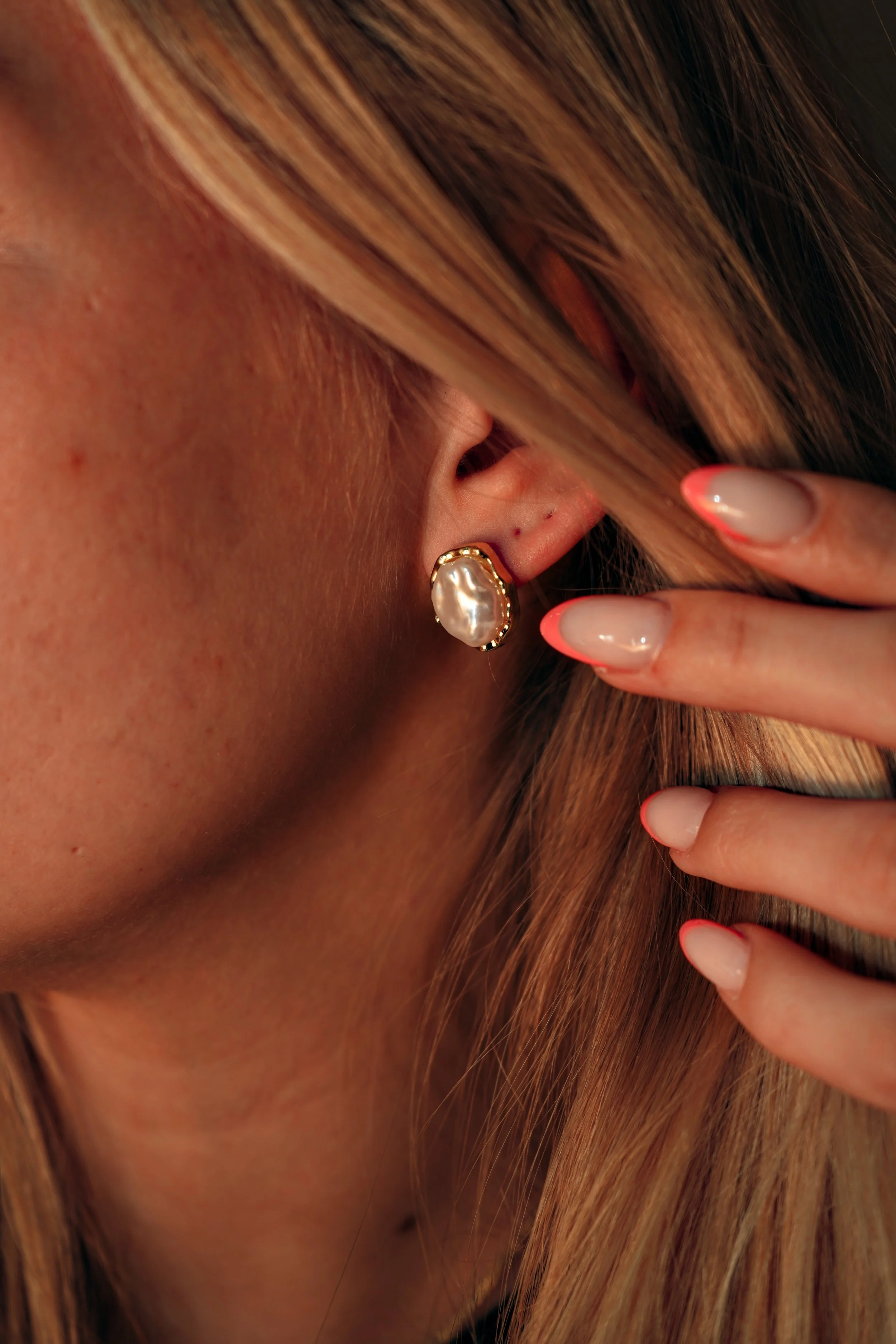 Tæt på øre med stor perlemorshalskædeørering, blond hår og velplejede negle.