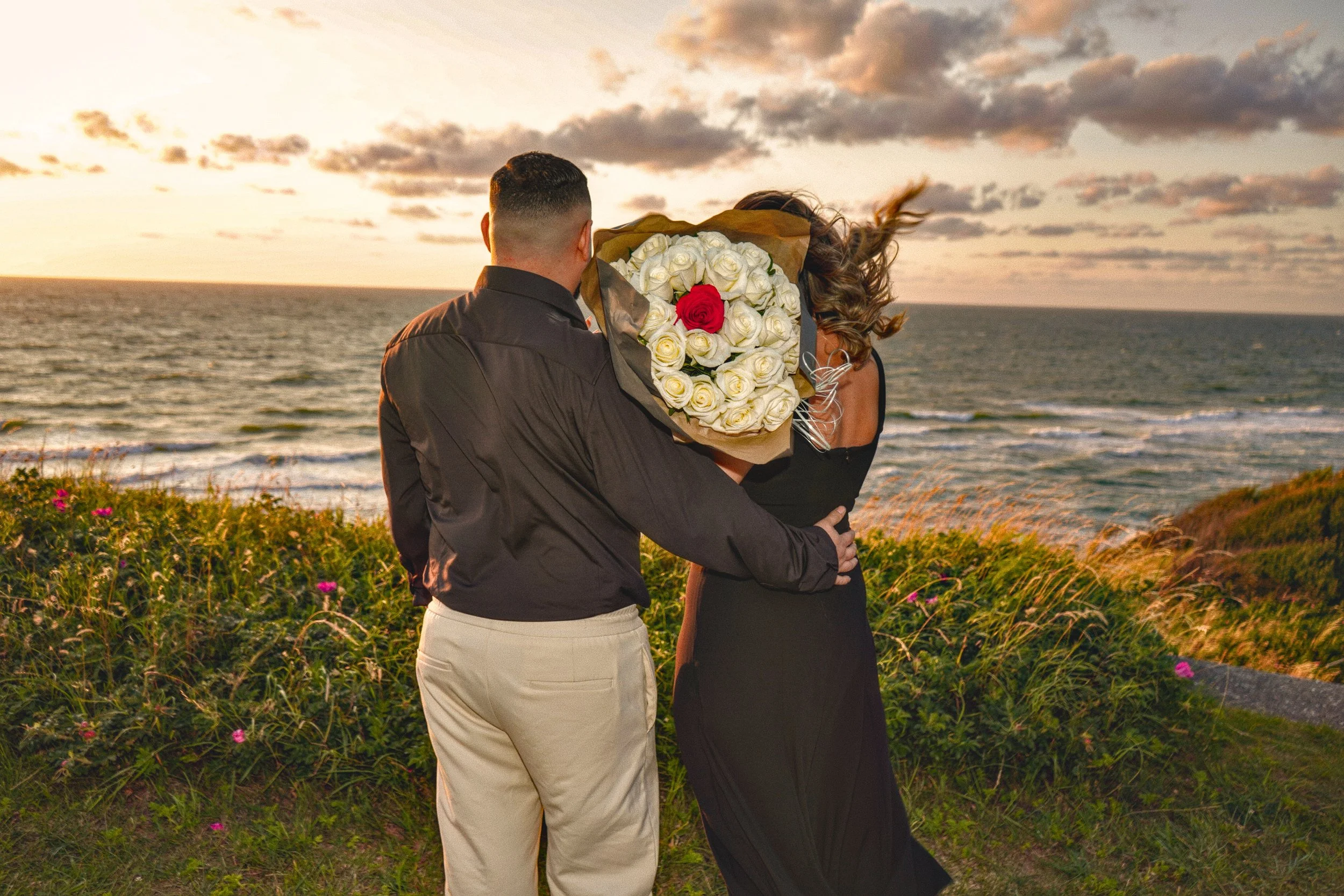 A couple standing on a grassy coastal cliff at sunset; the man is holding a large bouquet of white roses with a single red rose, and the woman is dressed in black with wind-blown hair, overlooking the ocean.