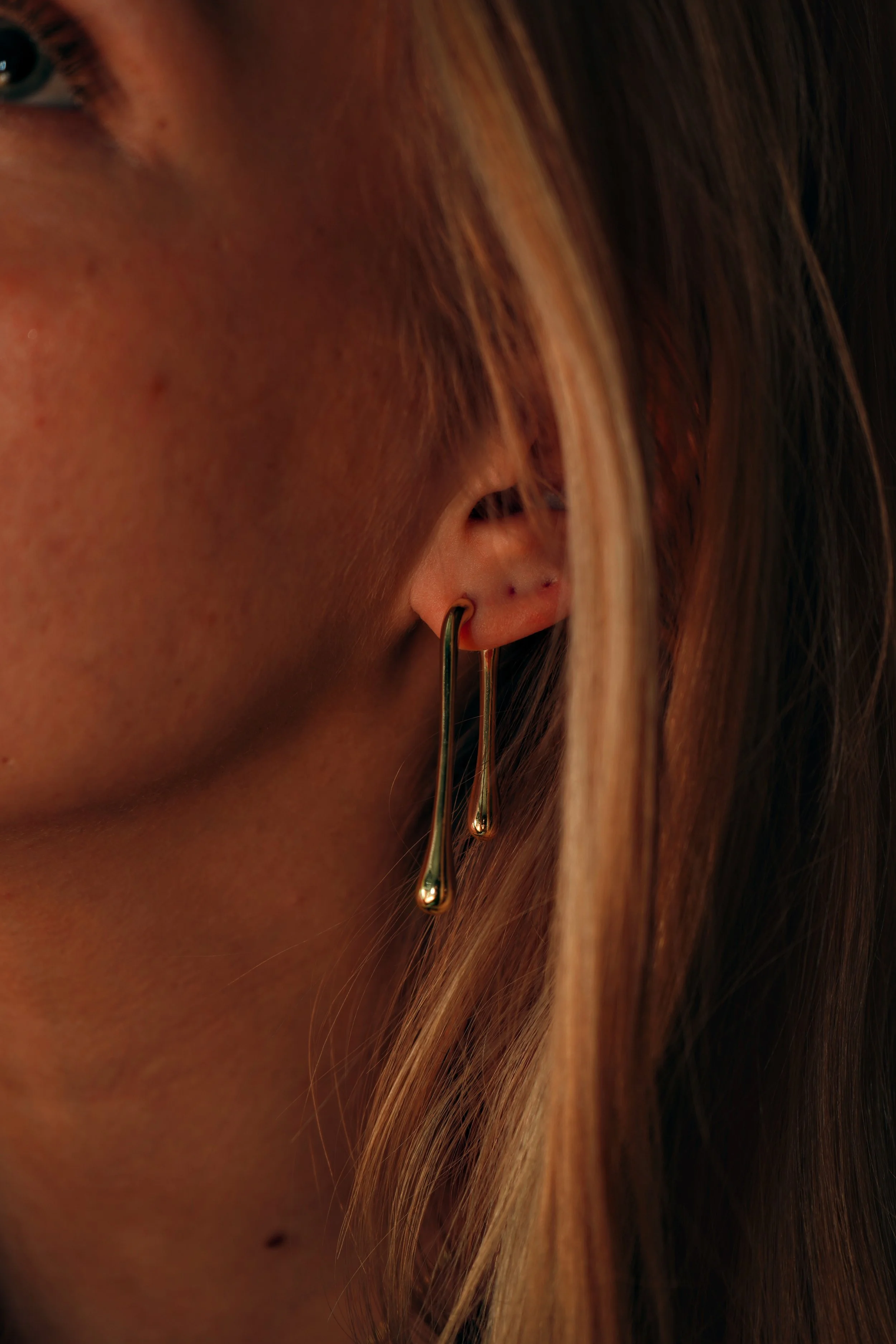 Nærbillede af en kvindes øre med lange guldøreringe, med hendes lange, lysebrune hår over den.