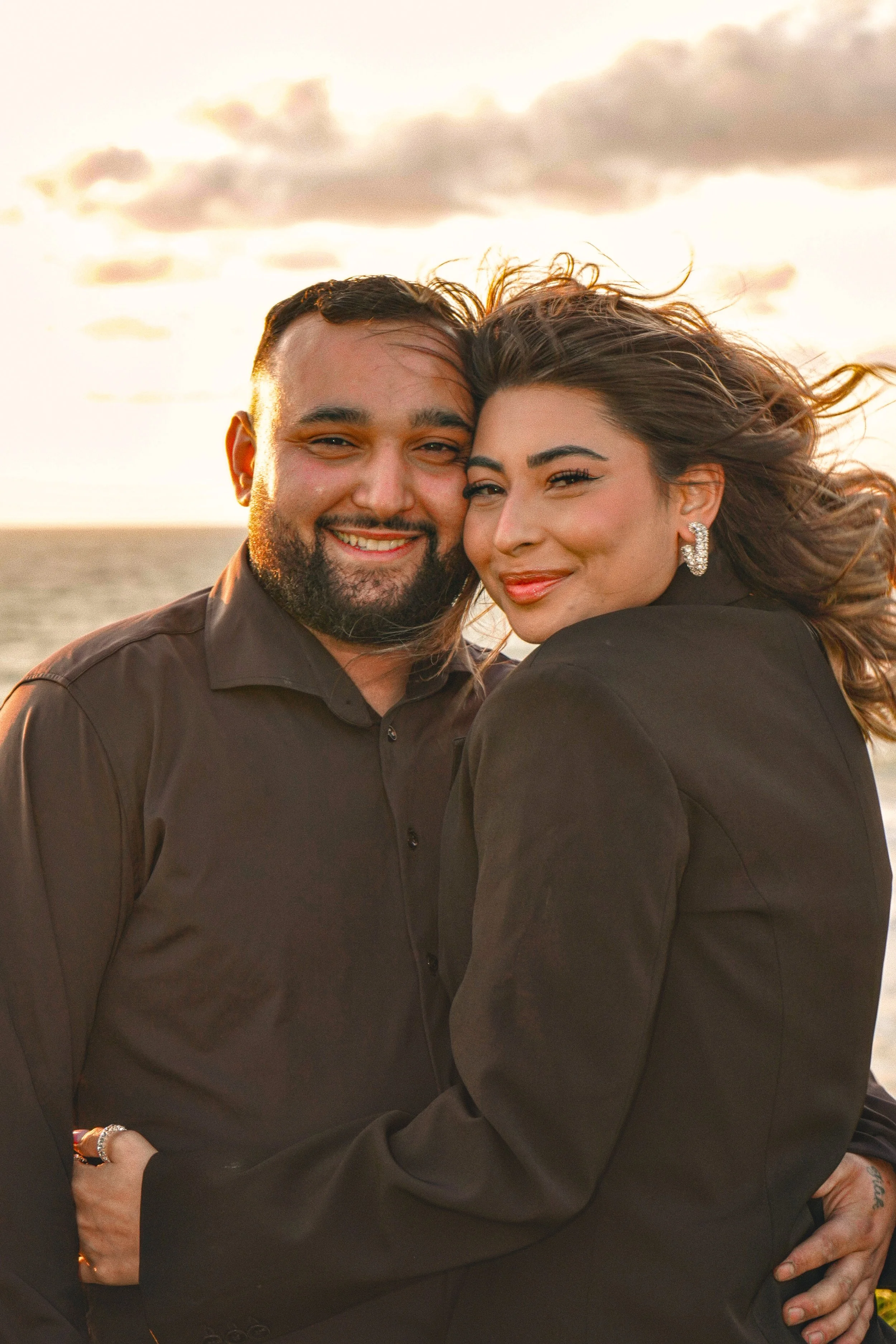 A happy couple smiling at the camera, standing close together at the beach during sunset, with ocean and cloudy sky in the background.