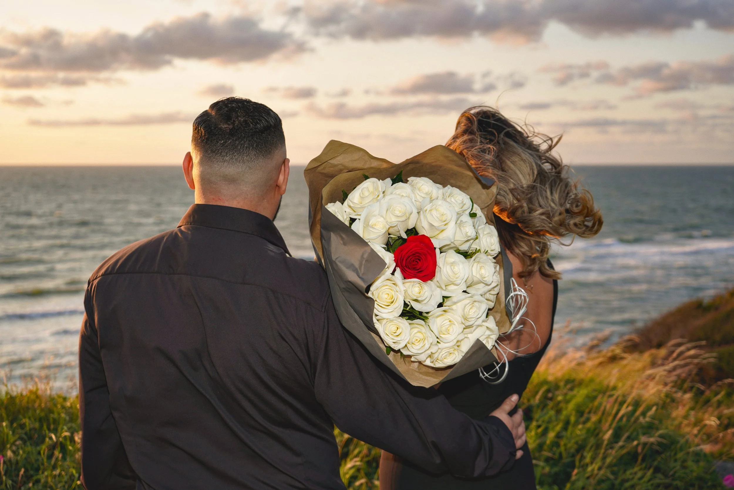A man is giving a woman a large bouquet of white roses with one red rose, near the ocean during sunset.