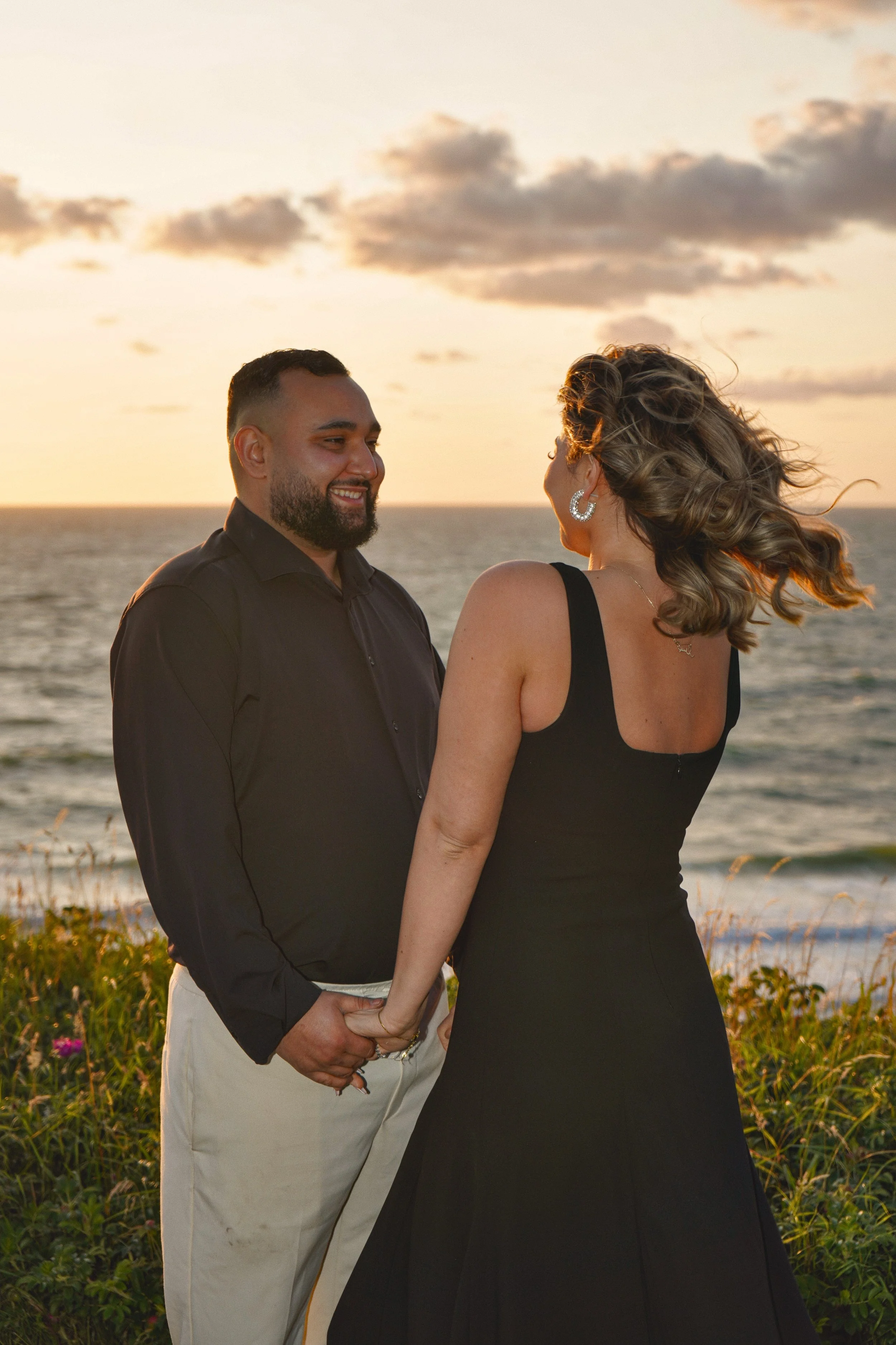 A couple holding hands, facing each other, on a beach at sunset. The man is dressed in a black shirt and white pants, and the woman in a black dress. The sky is orange and pink, with clouds and the ocean in the background.