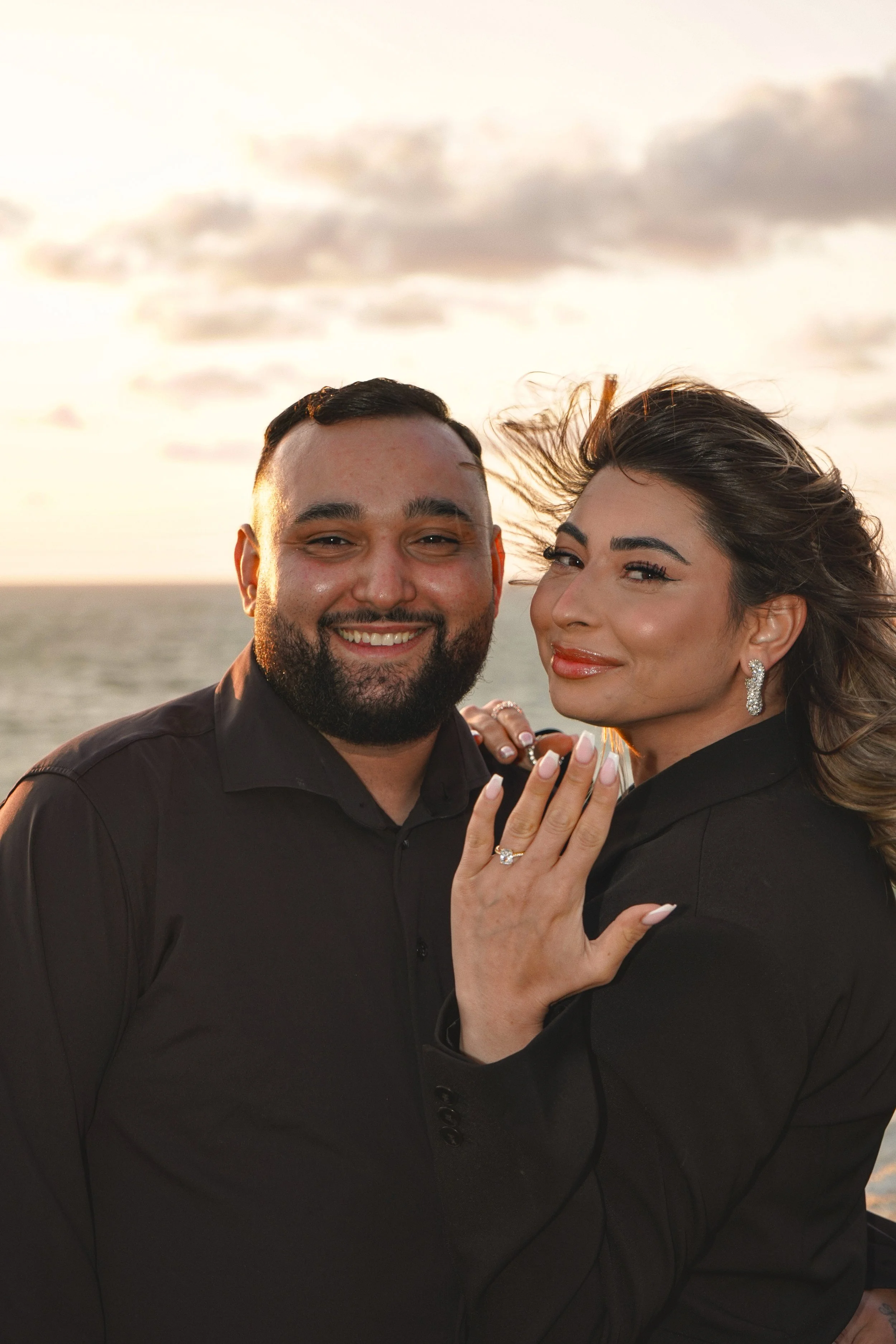 A happy couple showing off their engagement rings at sunset by the ocean.
