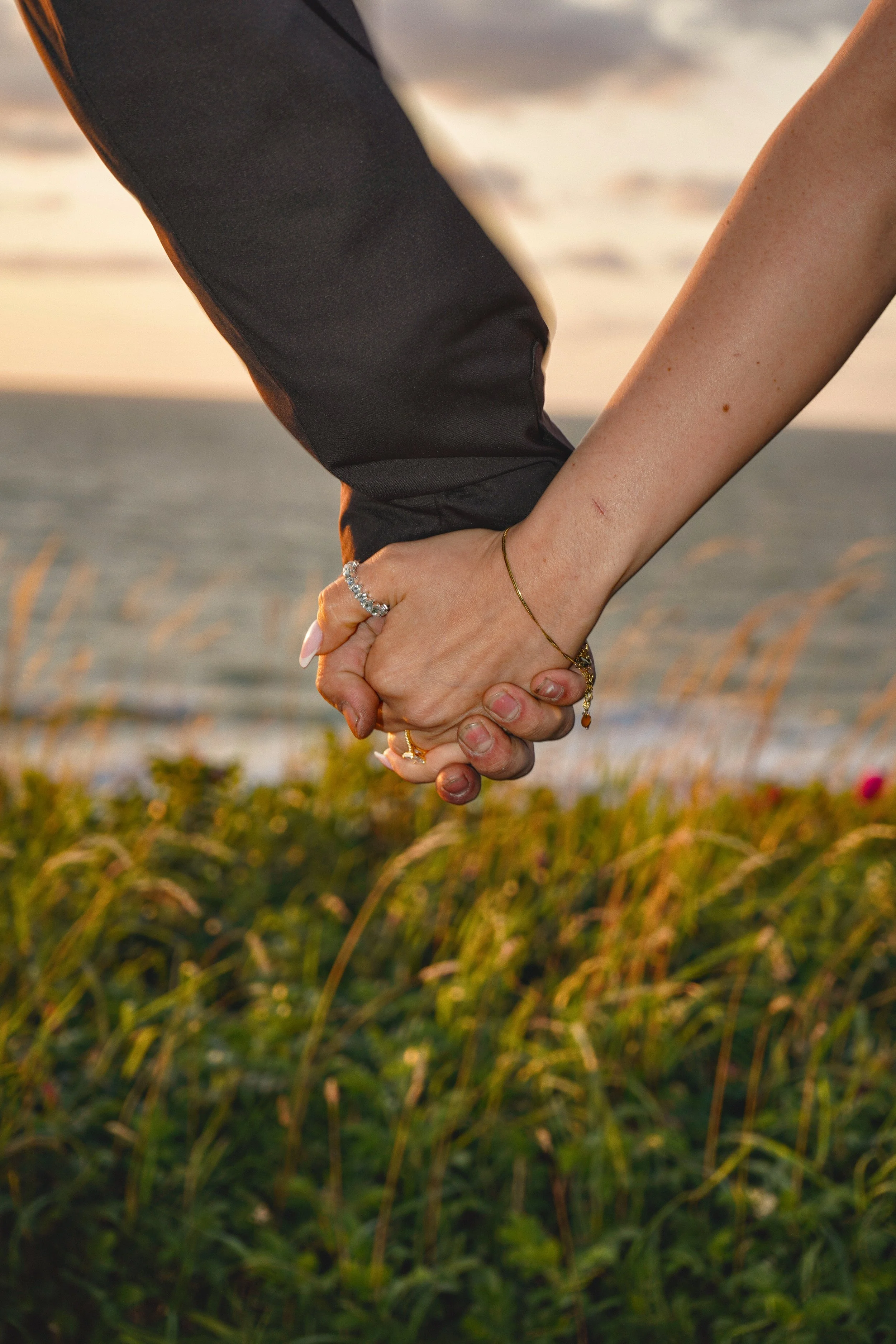 Close-up of a couple holding hands by the ocean during sunset, with the woman wearing rings and jewelry.