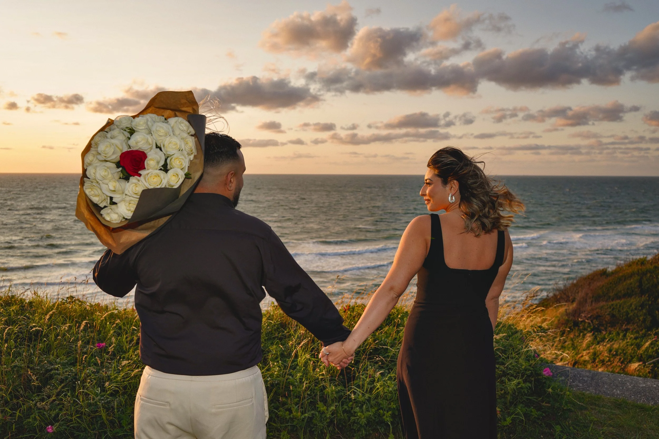 A couple holding hands by the ocean at sunset, with the man holding a bouquet of white roses and a red rose, the woman smiling and looking at the man, both dressed in elegant black and white clothing.