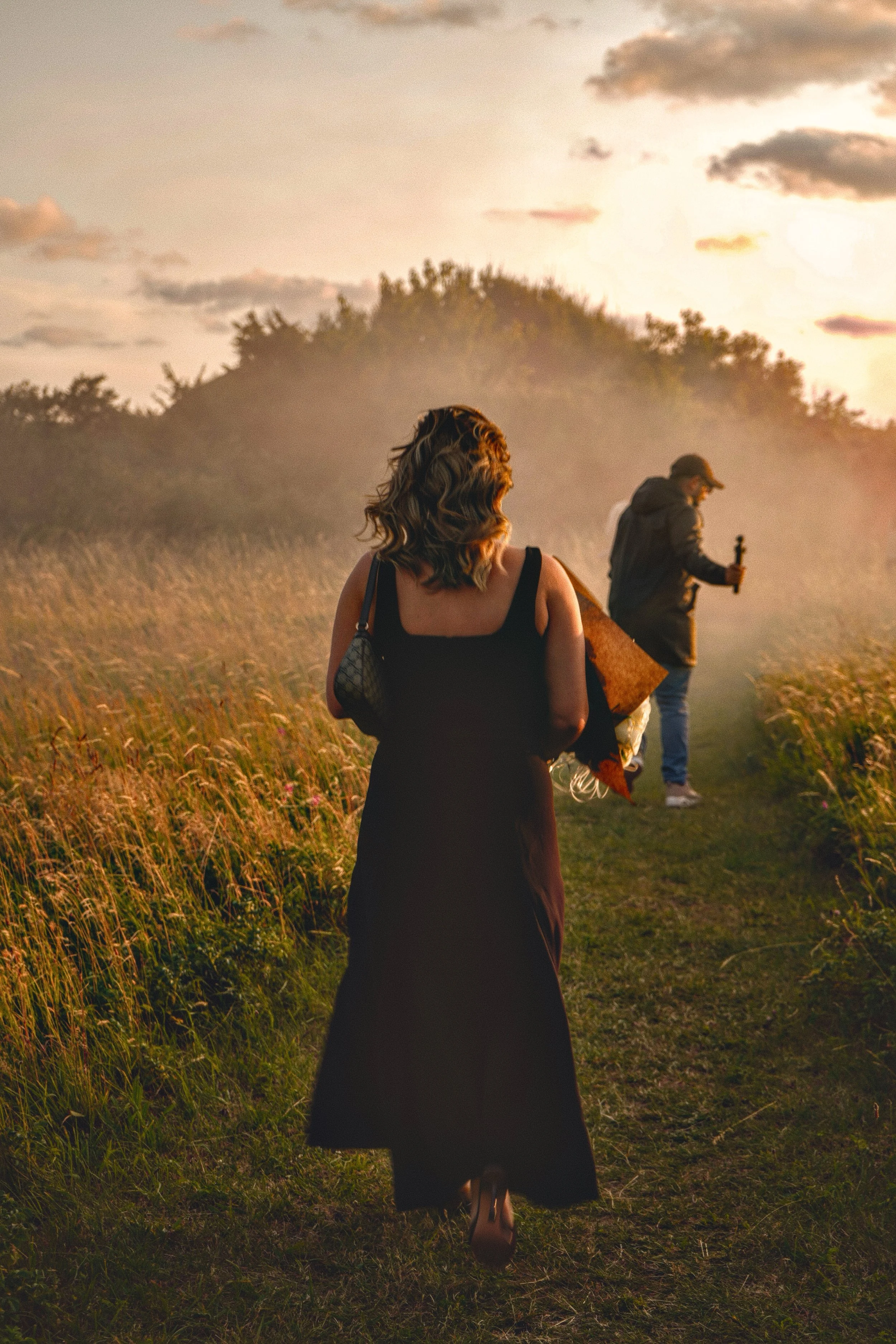 A woman with shoulder-length wavy hair wearing a black dress walking down a grassy trail at sunset, with a man in the background holding a camera or phone, surrounded by tall grass and trees.
