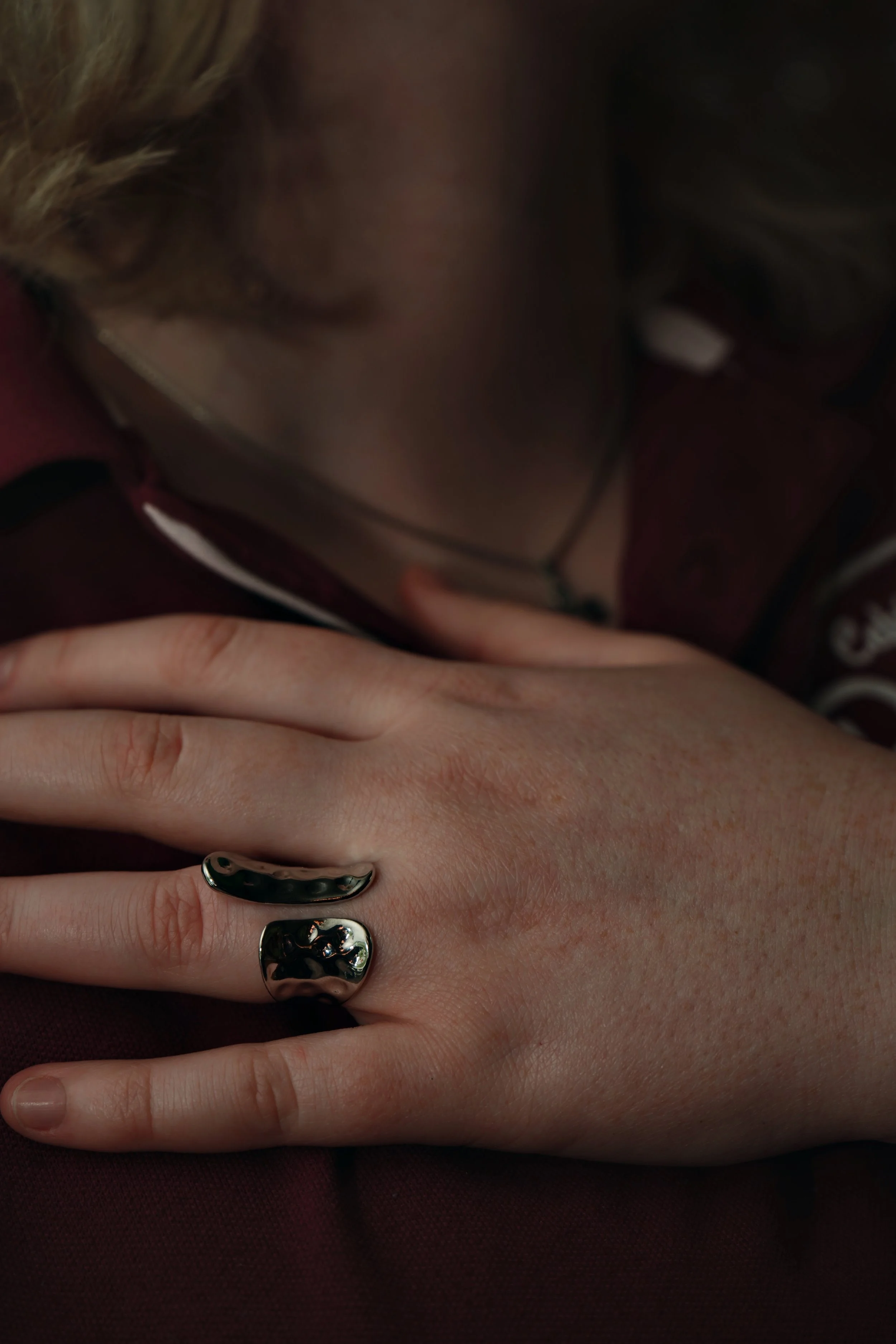 Nærbillede af en persons hånd med to store, glinsende sølvringe, liggende på en mørk overflade, med en person i baggrunden, der holder hånden.