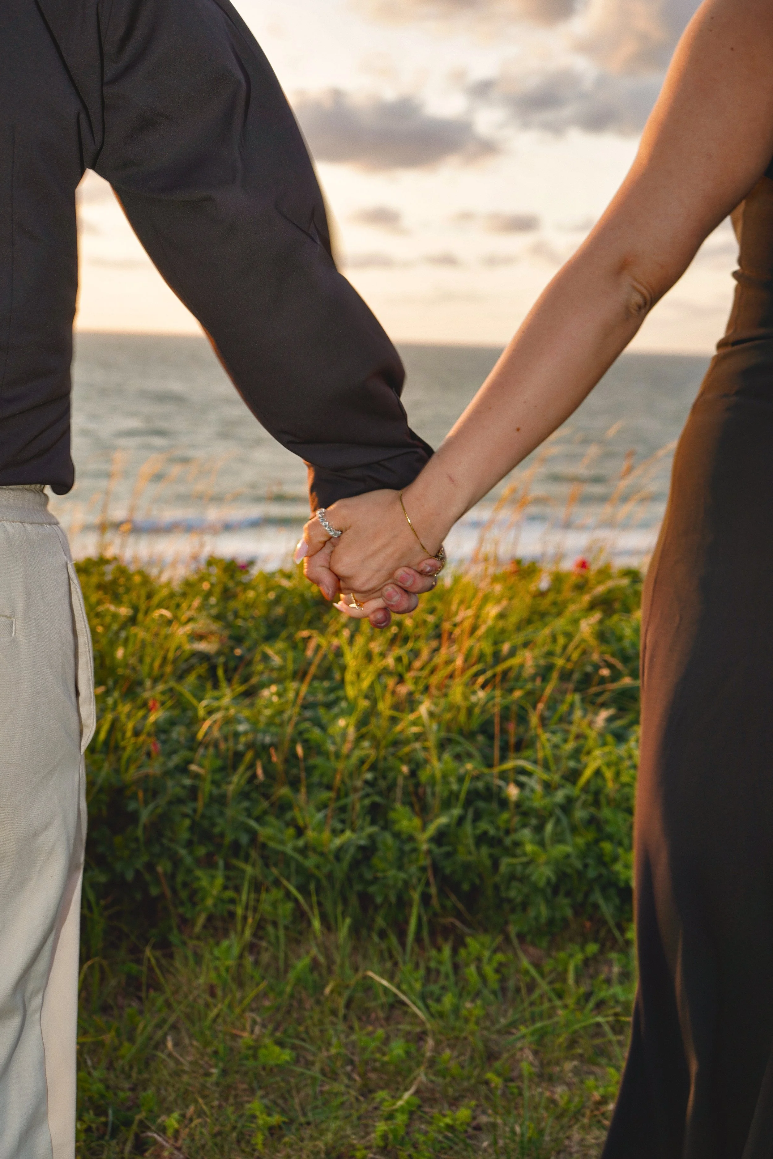 Close-up of a couple holding hands near the ocean at sunset, with grass and plants in the foreground.