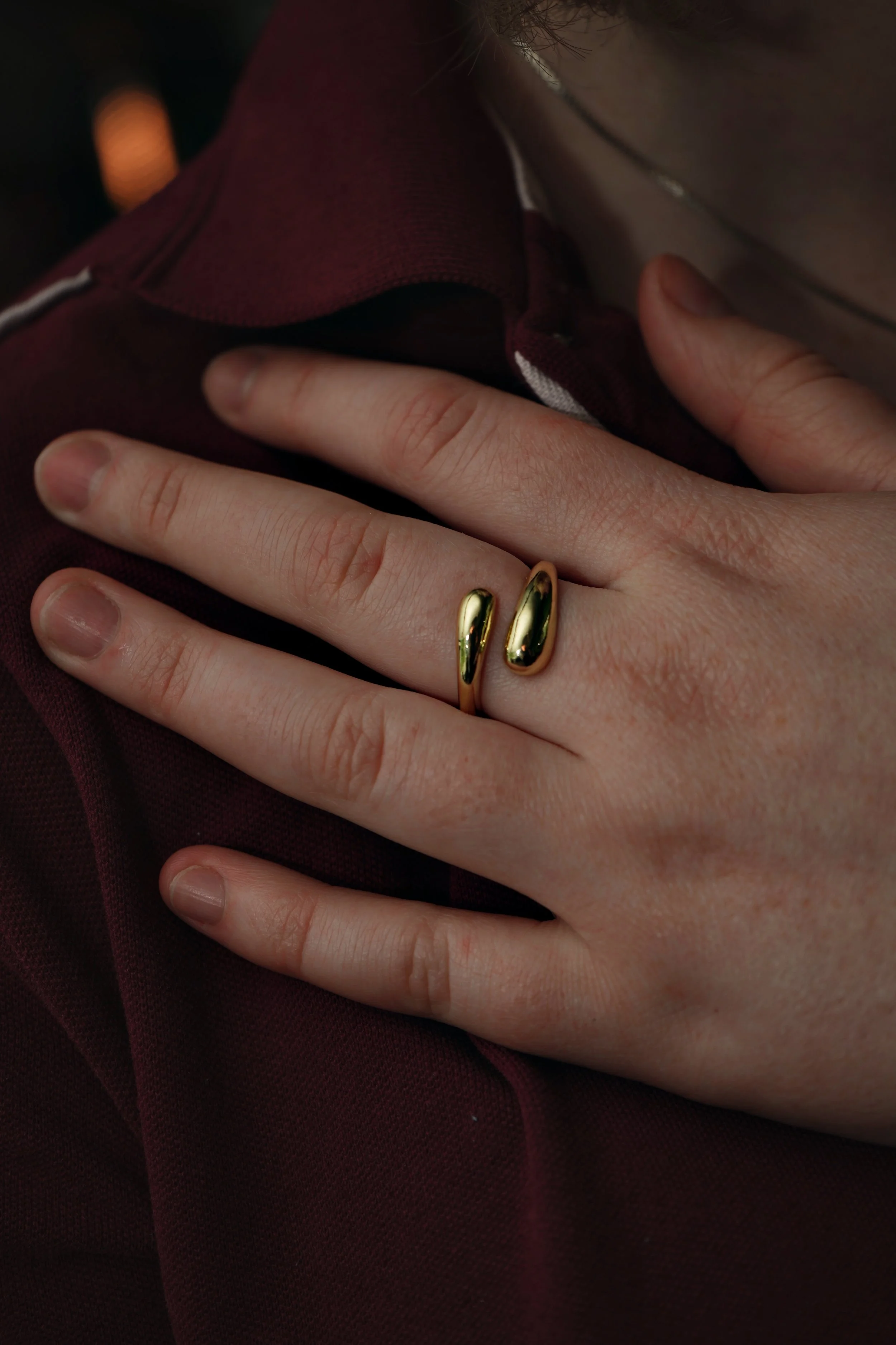 Nærbillede af en persons hånd, der viser en guldring på pegefingeren, mod en mørk rød skjorte.