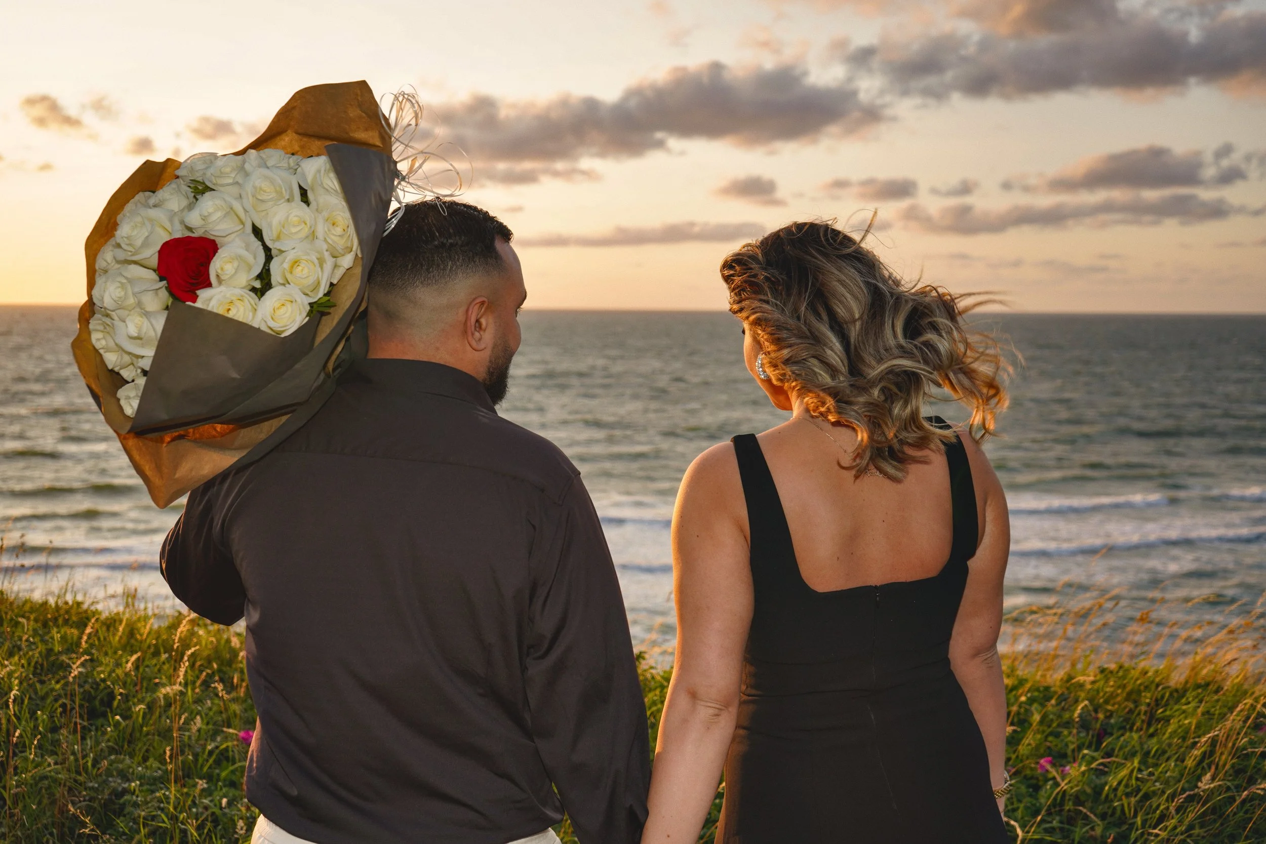 A couple stands facing the ocean at sunset, the man holding a large bouquet of white roses with a single red rose on top, both with their backs to the camera.