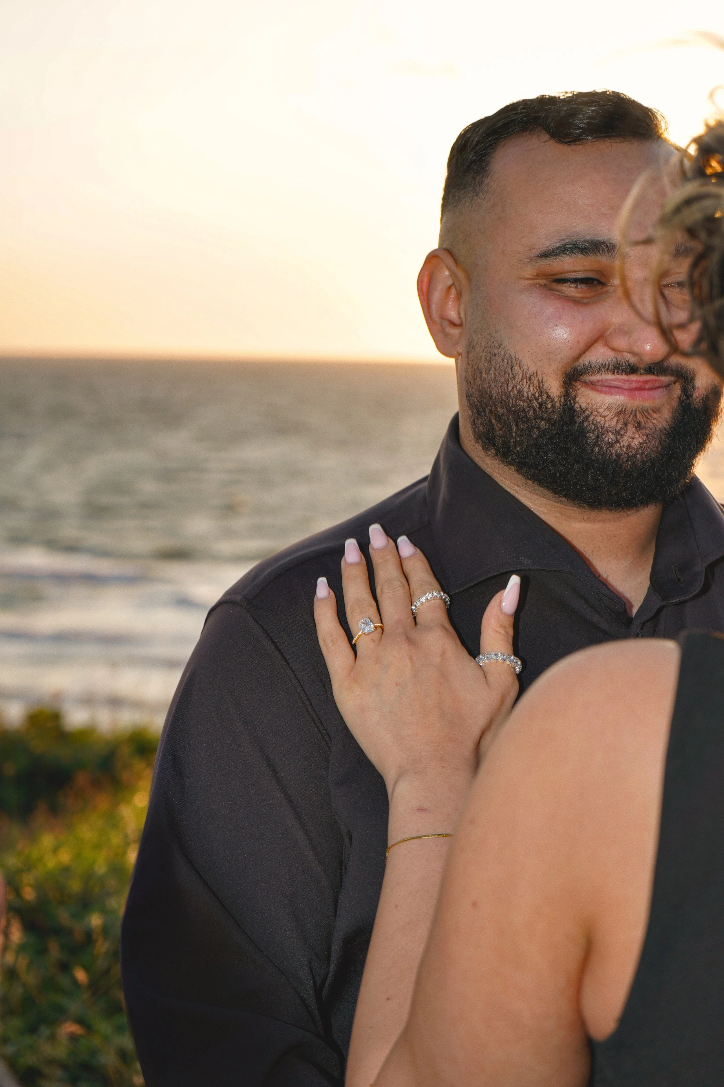 A couple at the beach during sunset, with the woman showing rings on her fingers and the man smiling.
