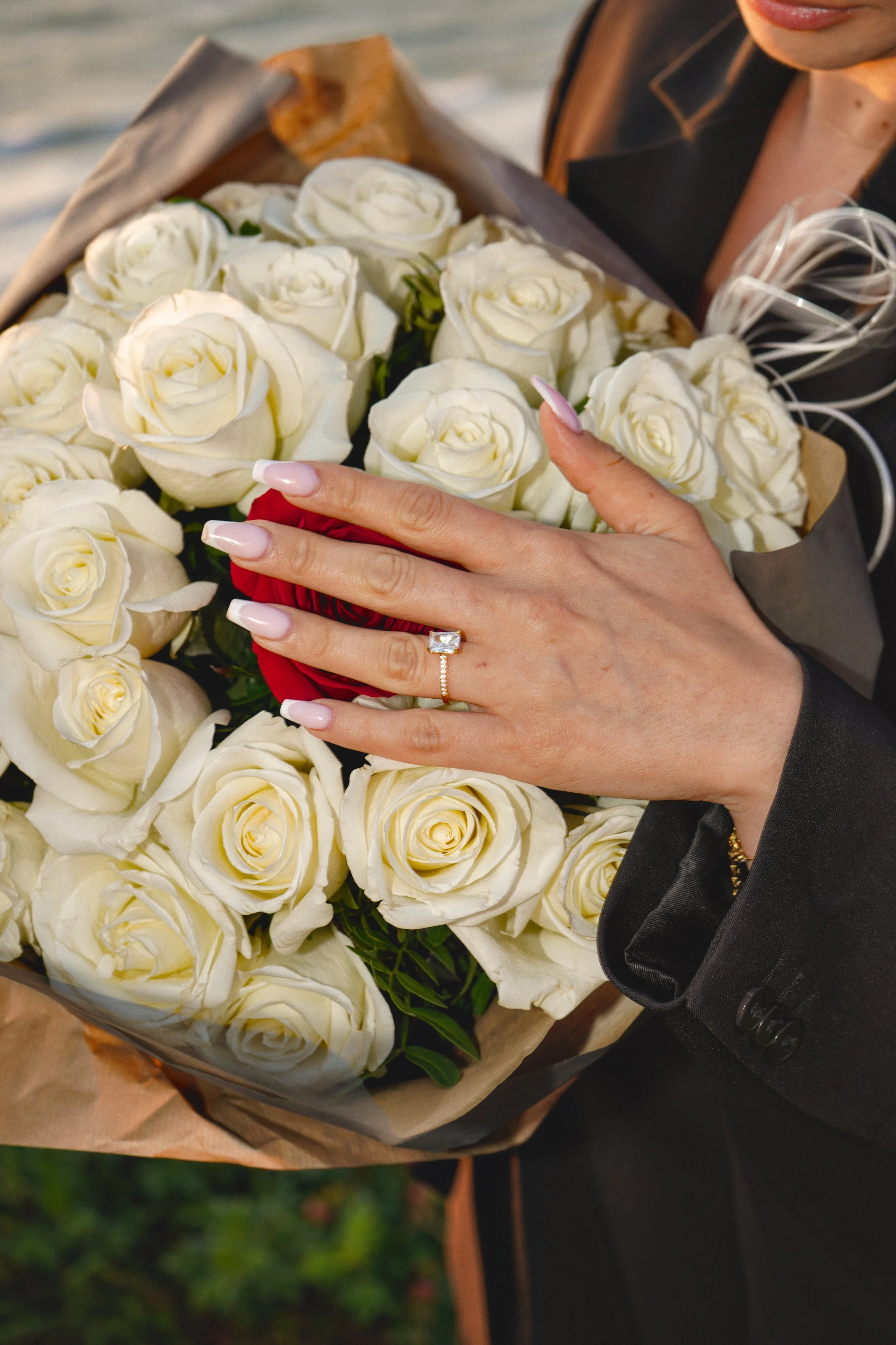 A woman displaying a ring on her finger while holding a bouquet of white roses and one red rose, possibly celebrating an engagement or special occasion.