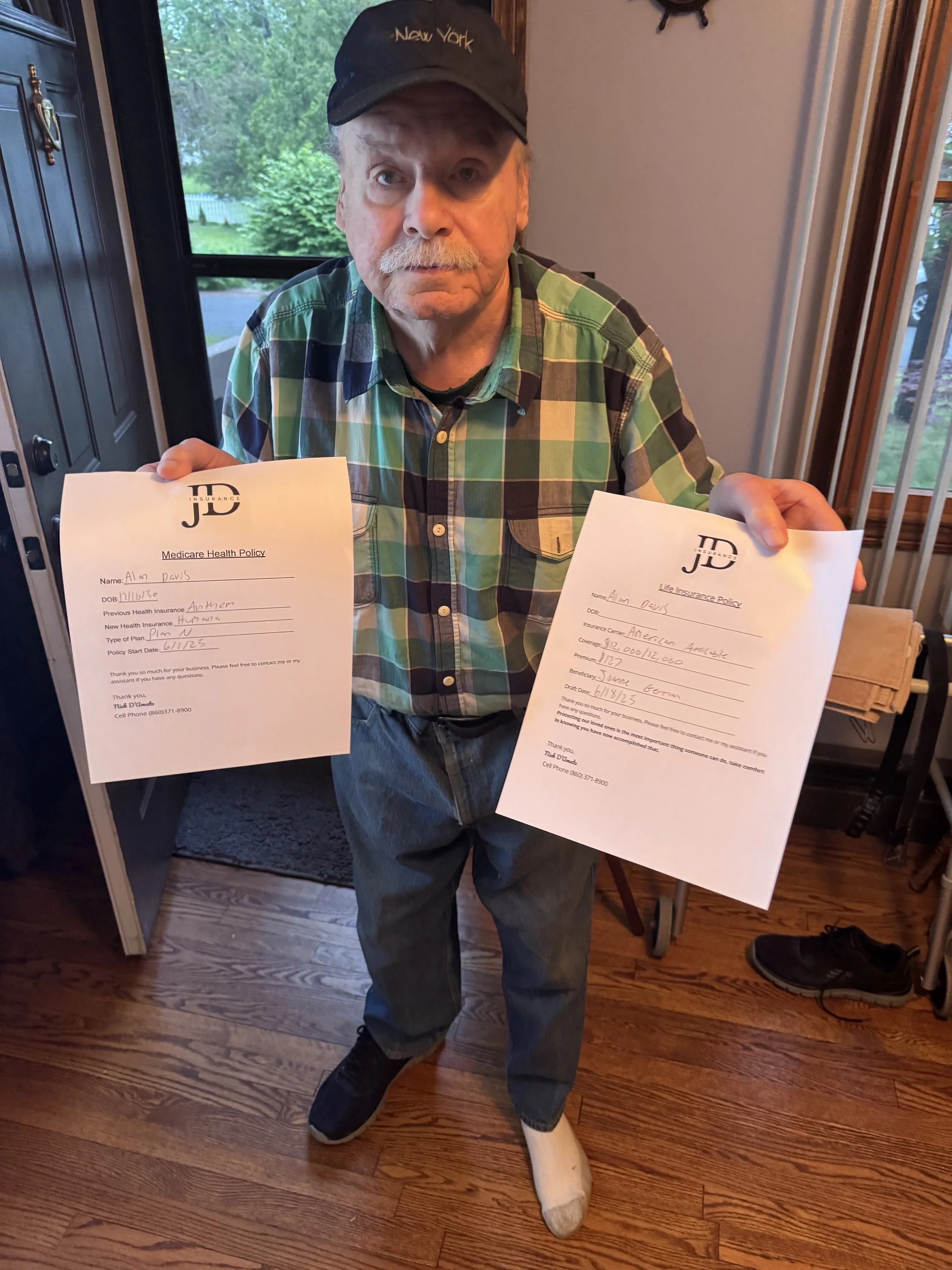 An elderly man wearing a cap, checkered shirt, and jeans, holding two documents, one in each hand, inside a home near a window with a view of greenery outside.