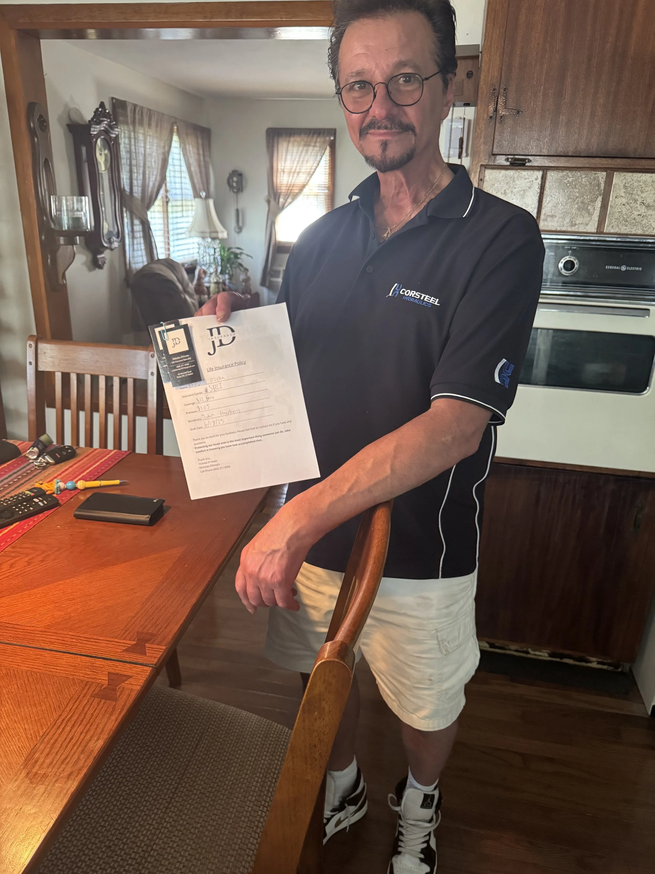 A man standing in a kitchen holding a document, wearing a navy blue polo shirt, khaki shorts, and sneakers, with glasses and facial hair. The kitchen has wooden furniture and a living room with curtains and lamps in the background.