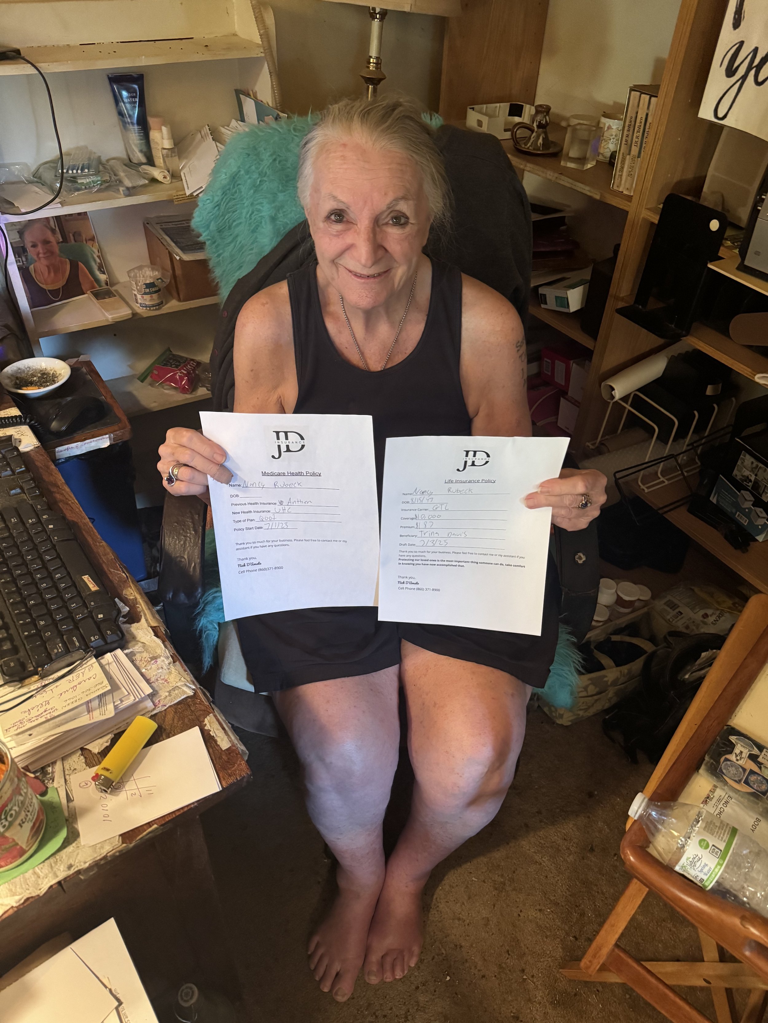 An elderly woman sitting on a chair in a cluttered room, holding two documents, smiling at the camera.