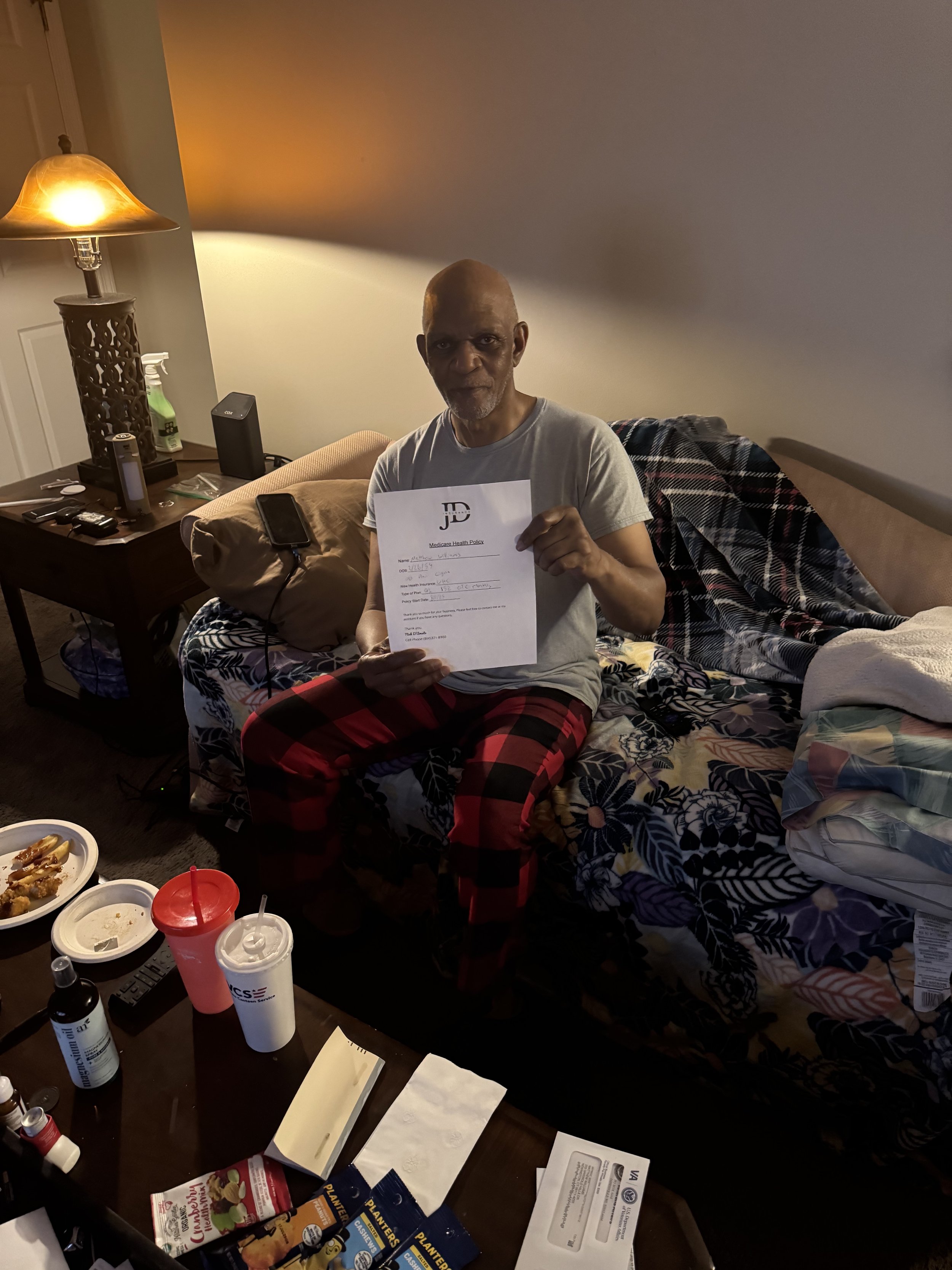 An elderly man sitting on a floral bedspread holding a document in a dimly lit room with a table of food and drink in front of him.