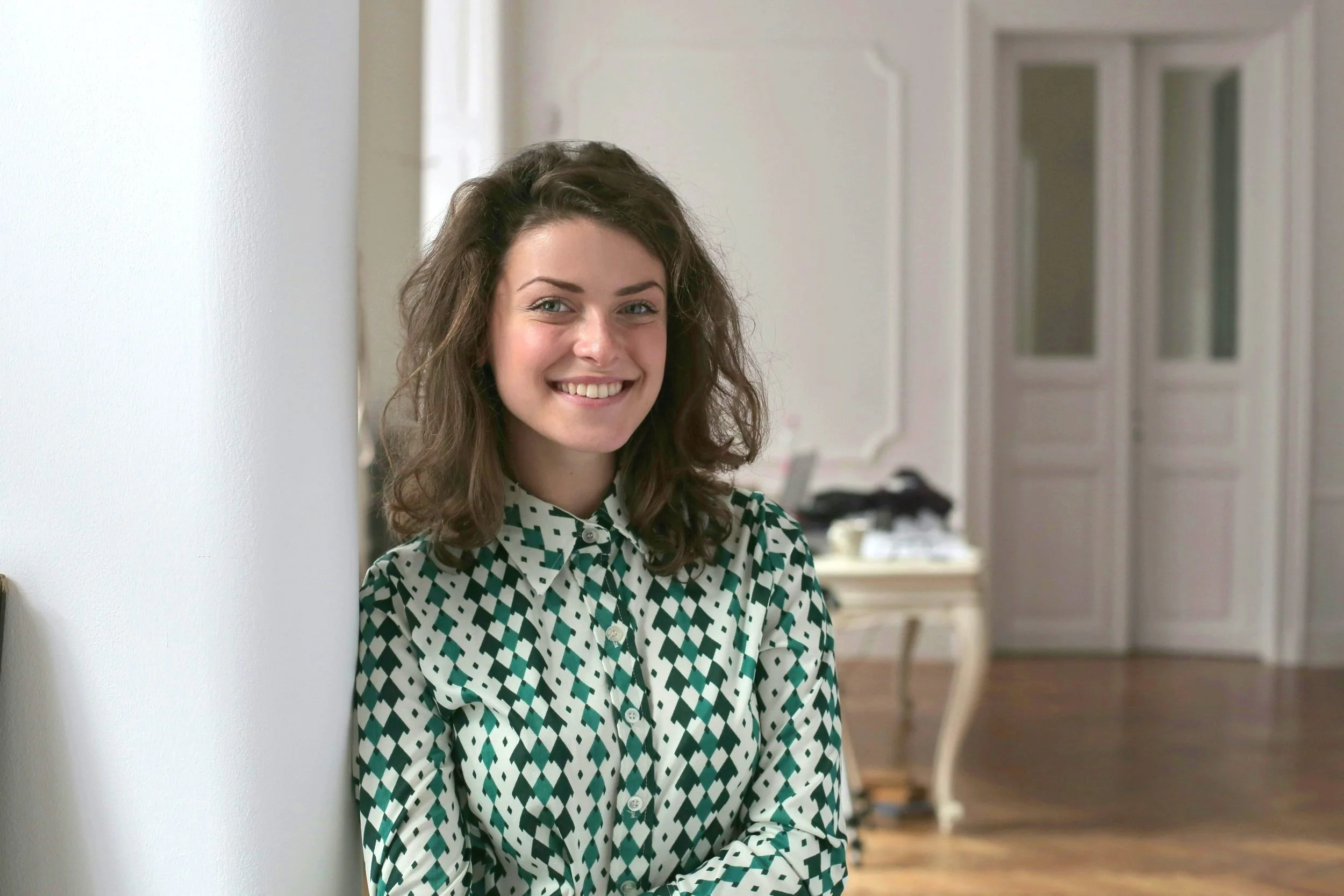 A woman with wavy brown hair, wearing a black and white geometric patterned blouse, smiling while leaning against a white wall in a well-lit room with a blurred background of a room with high ceilings and glass-paneled doors.