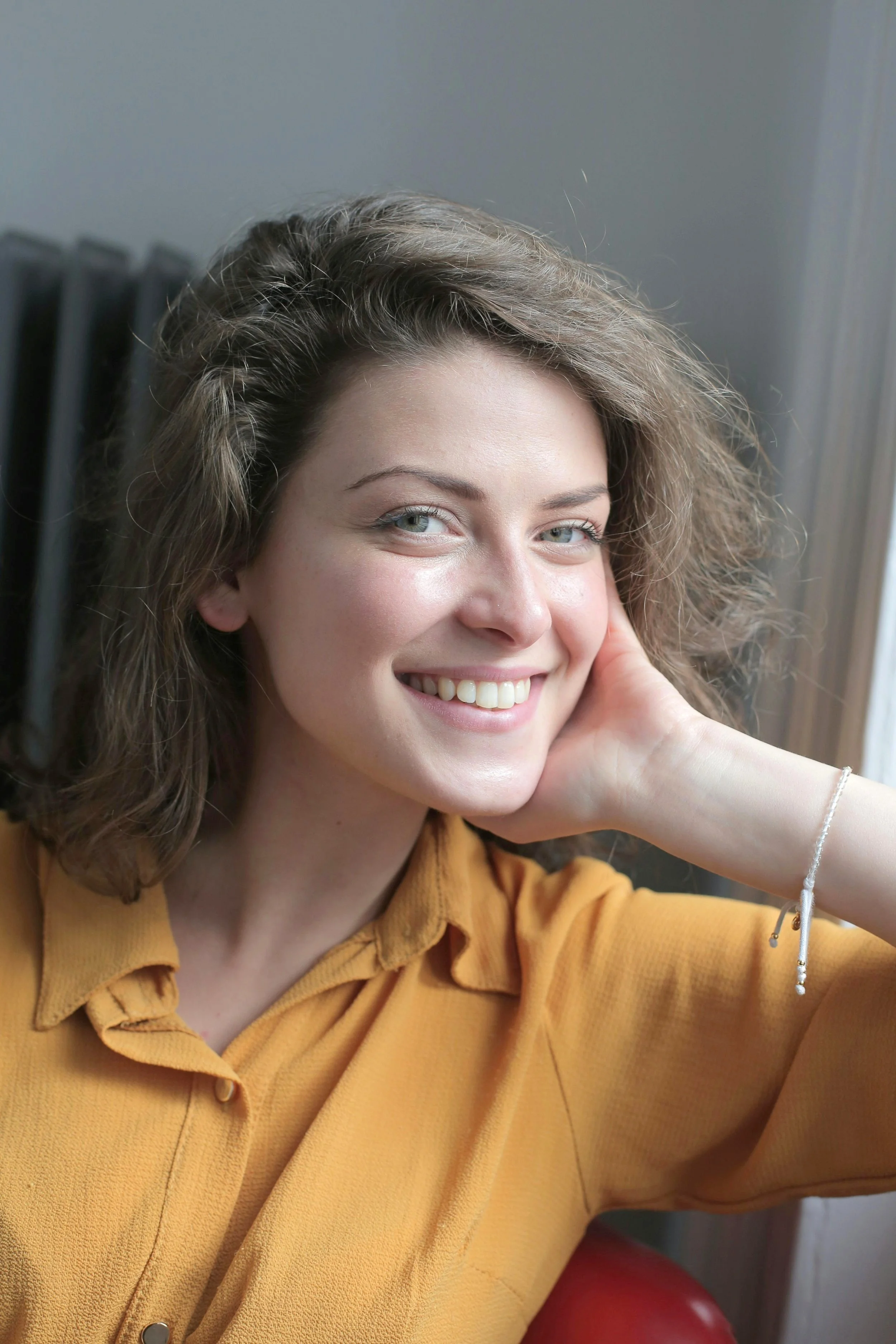 A woman smiling, with light brown hair and wearing a mustard yellow shirt, touching her face with her right hand, in front of a window and radiator.