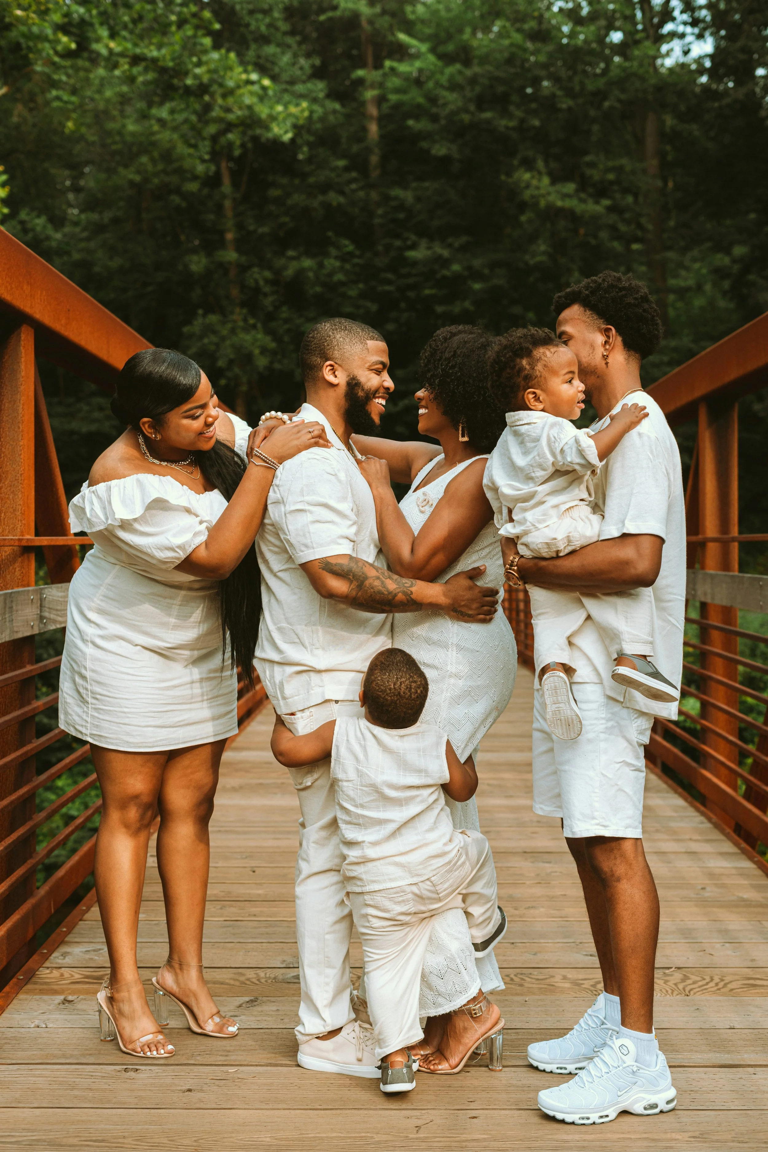 Family of six of African American descent on a wooden bridge surrounded by green trees, smiling and hugging each other, wearing beige and white outfits, with two children and all sharing joyful moments.