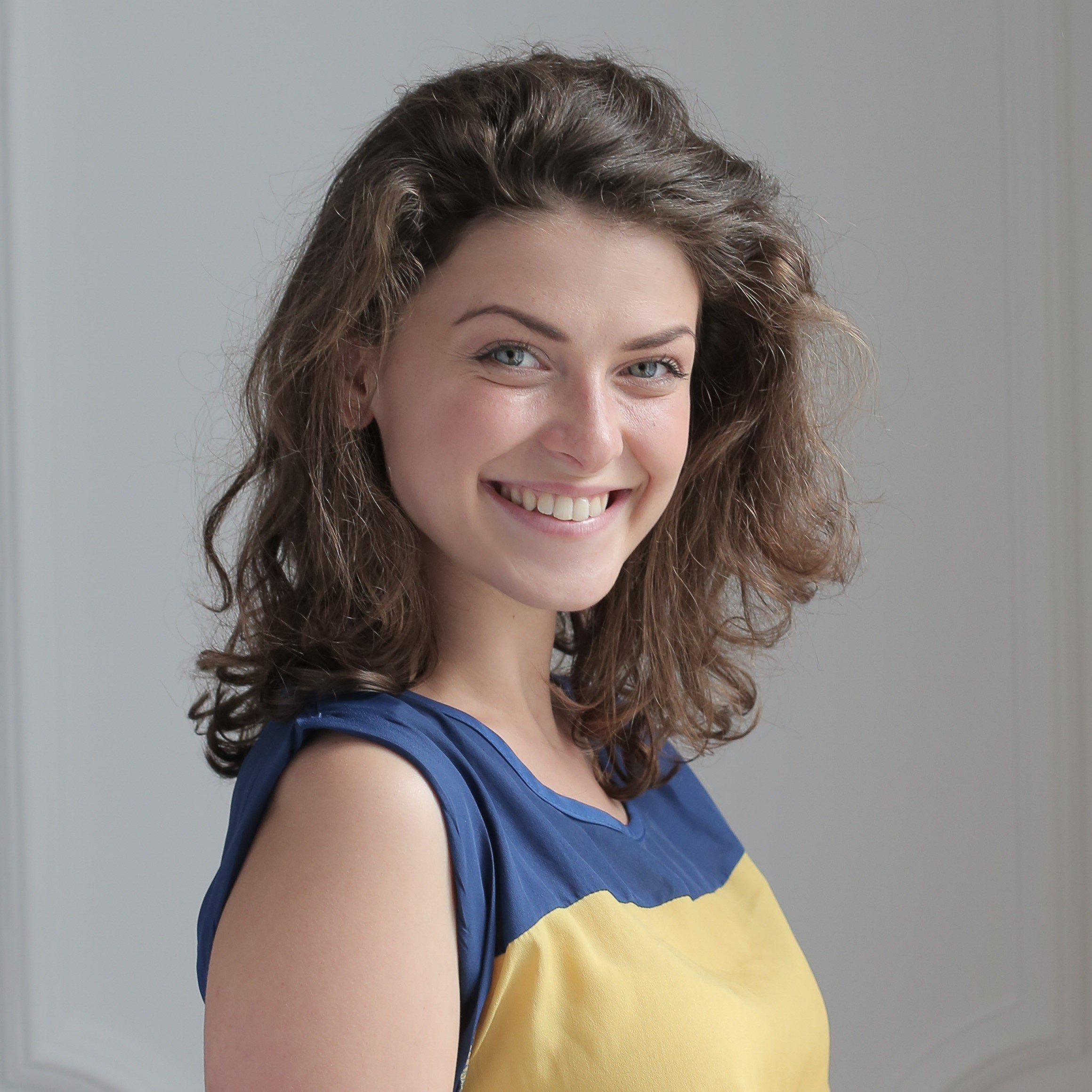 Smiling woman with curly brown hair and blue eyes wearing a blue and yellow sleeveless dress, standing in front of a plain gray wall.