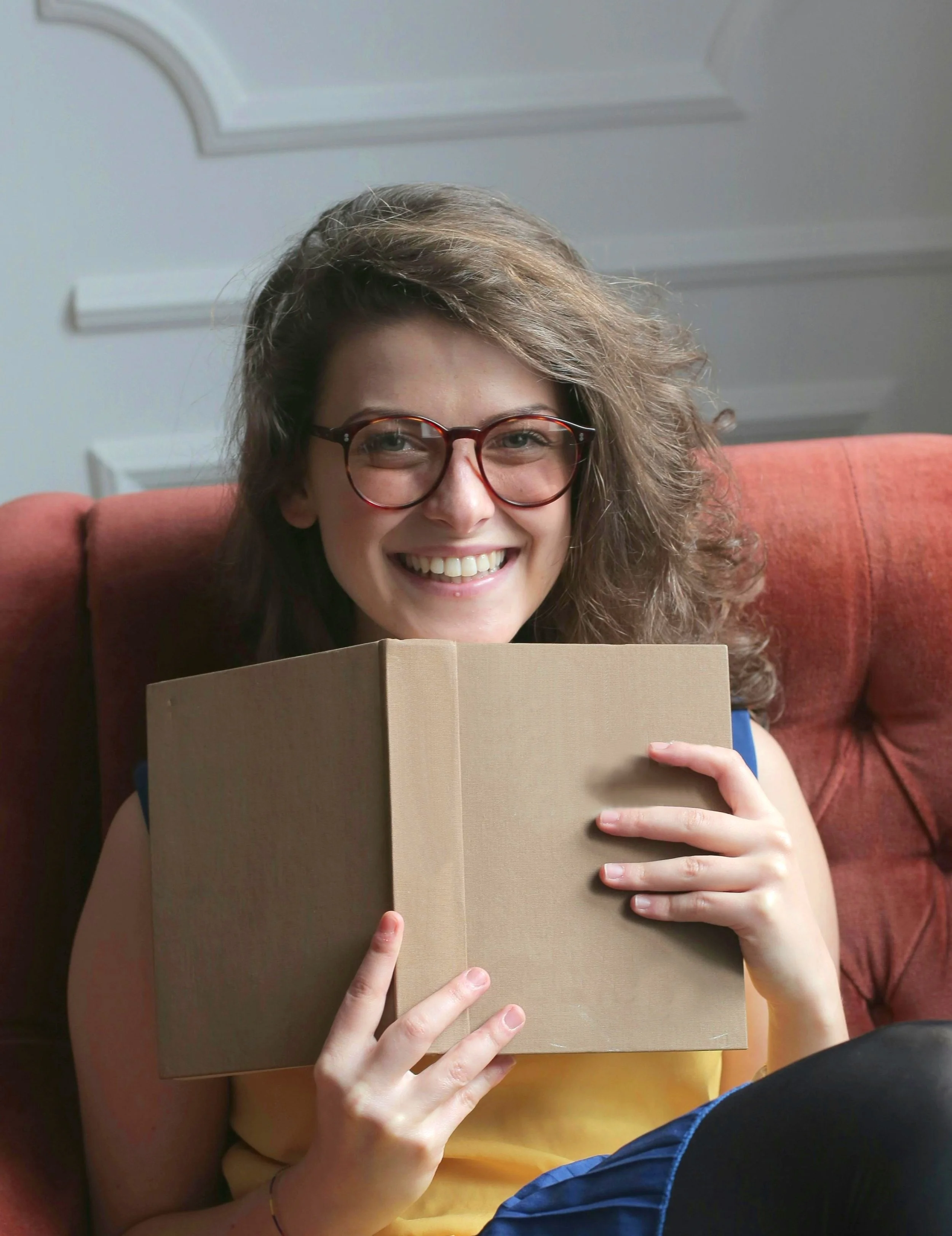 A person with glasses holding a closed beige book in front of their face while sitting on a red cushioned seat. They are smiling and wearing a yellow top.