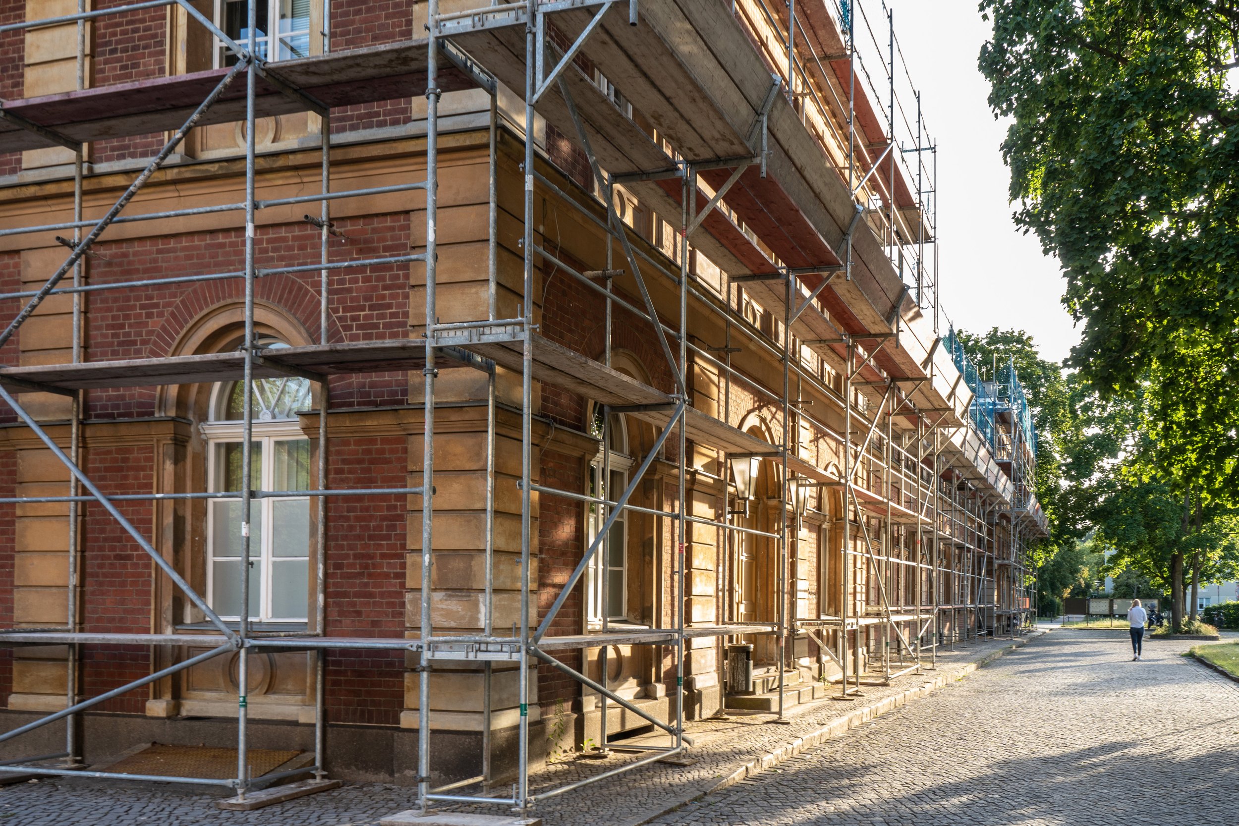 A historic brick building undergoing renovation with scaffolding along the facade, set in a tree-lined street during late afternoon.