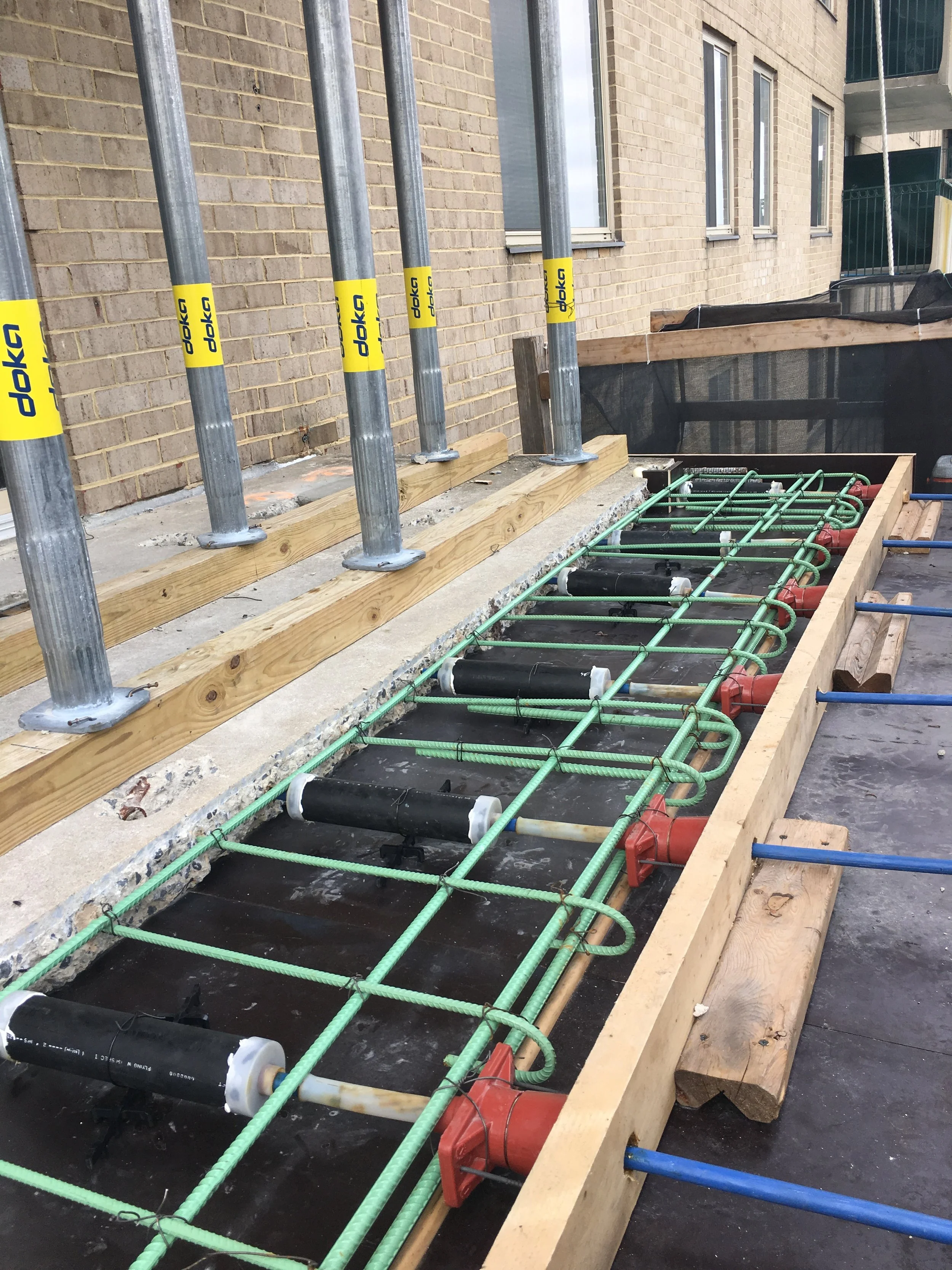 Construction site with plumbing installation, green pipes, black foam rollers, and wooden framing next to a brick building.