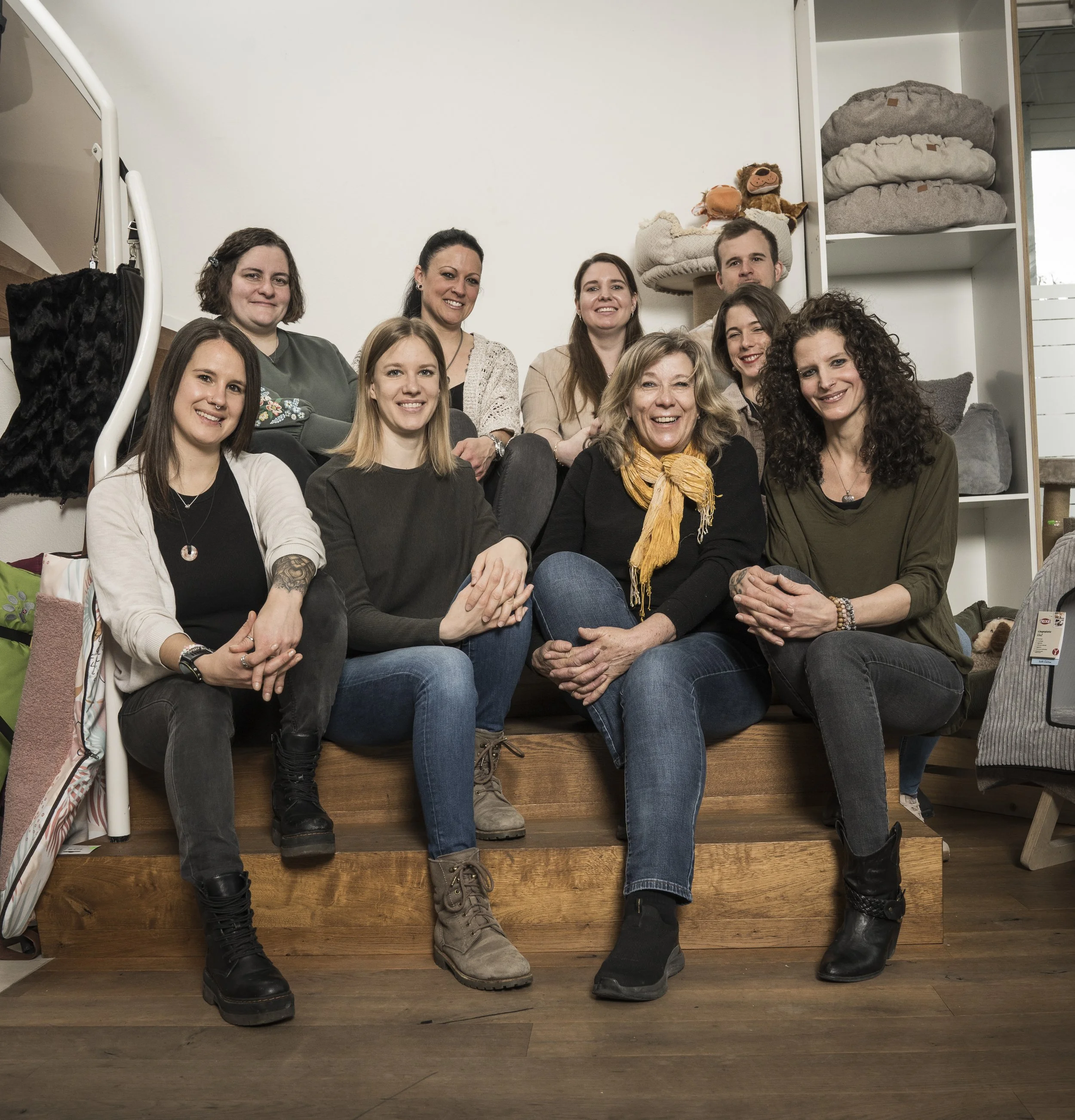 Gruppe von zehn Menschen sitzt gemeinsam auf Holzstufen in einem Raum mit weißen Wänden und Regalen, die Decken- und Kissenstapel sowie Plüschtieren enthalten.