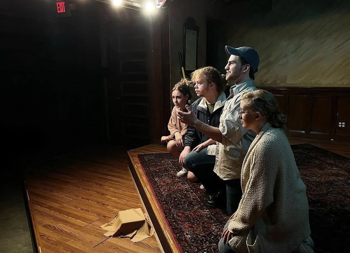 Four people kneeling on a stage in a dark room with one person speaking.