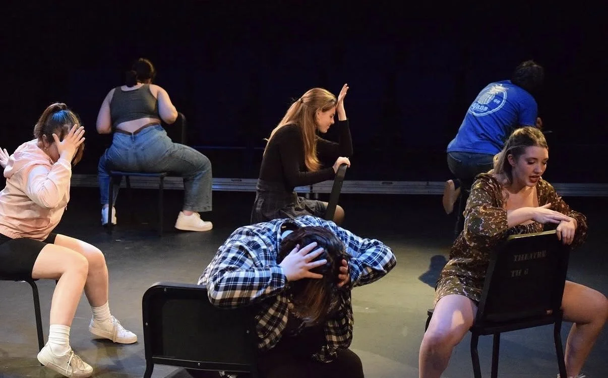 Six women sit on chairs on a dark stage, each holding their head or face in distress in a theatrical performance or rehearsal.