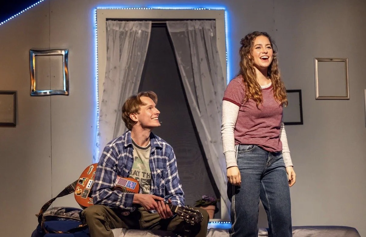 A young man sits on a bed holding an acoustic guitar, while a young woman stands next to him, smiling and laughing. The room has gray walls, framed pictures, a window with curtains, and a neon blue light border.