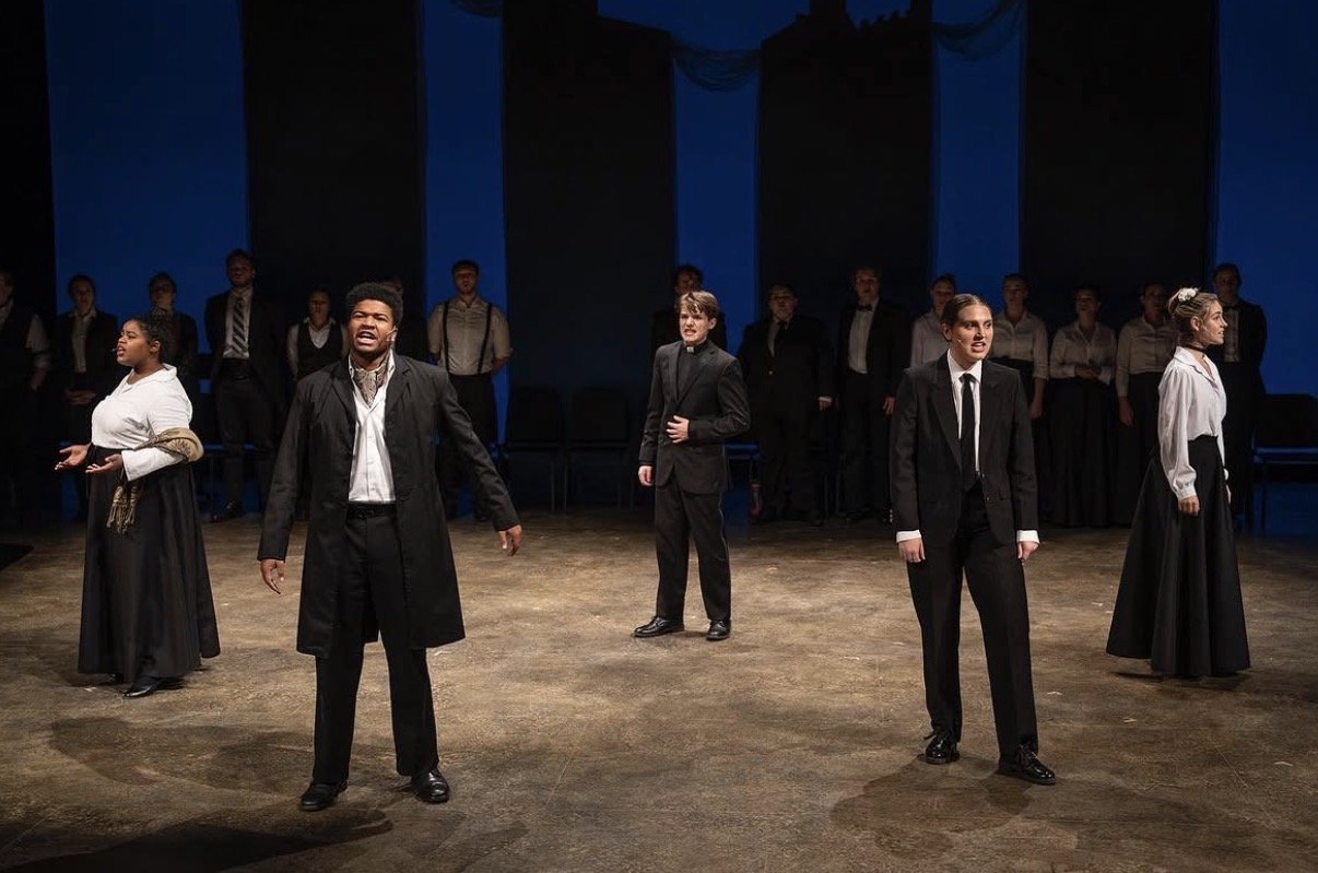 The scene shows a theatrical performance with actors on-stage, dressed in formal and period costumes, with some acting passionately and others standing in the background.