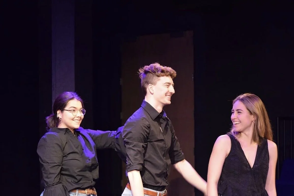 Three young actors on stage during a performance, smiling and holding hands, with a black background.