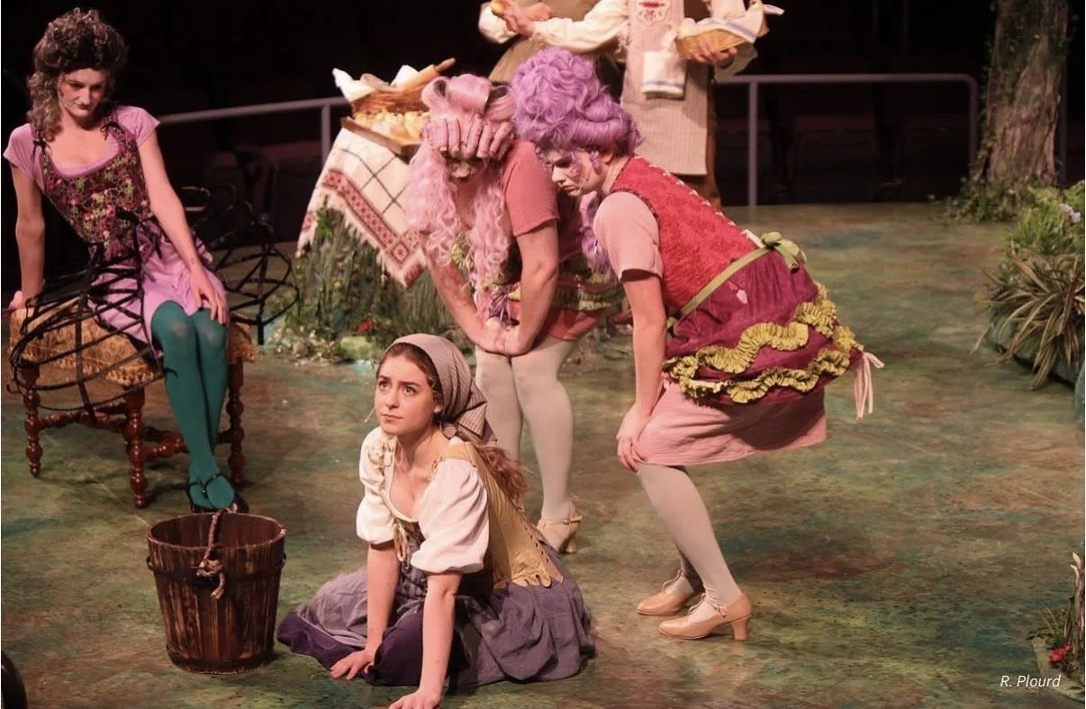 Women dressed as characters from a stage play, with a young girl sitting on the stage floor surrounded by plants and props.
