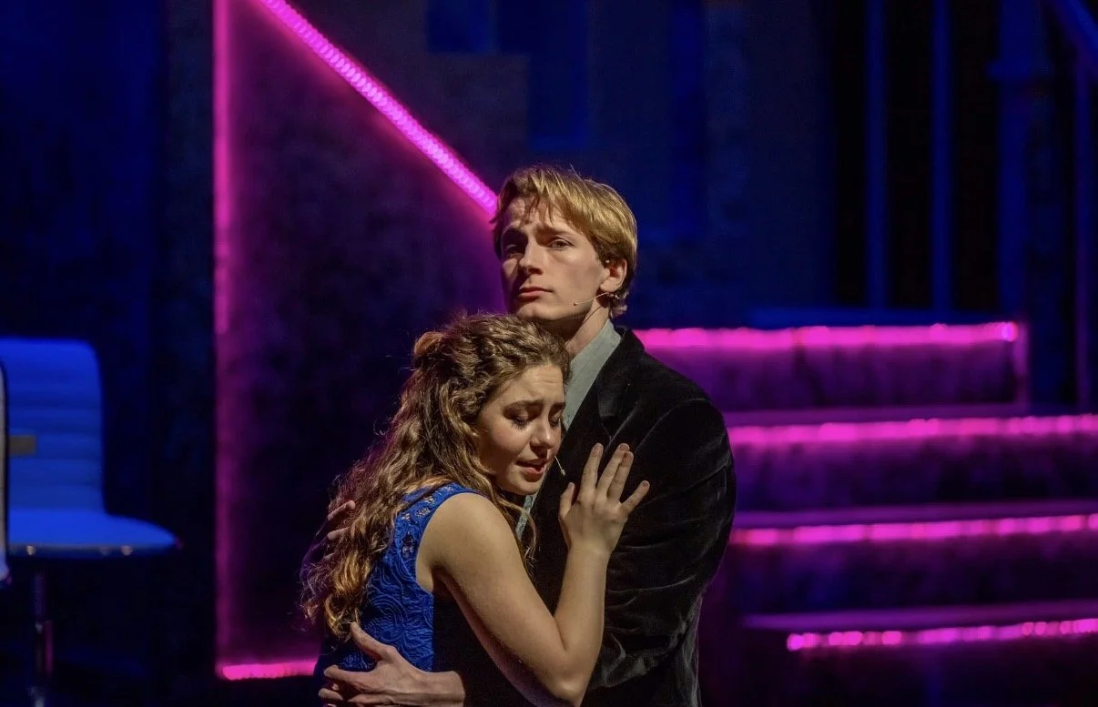 A man and woman share an emotional moment on stage; the woman appears sad and is holding onto the man, who looks serious, with neon pink and blue lighting in the background.