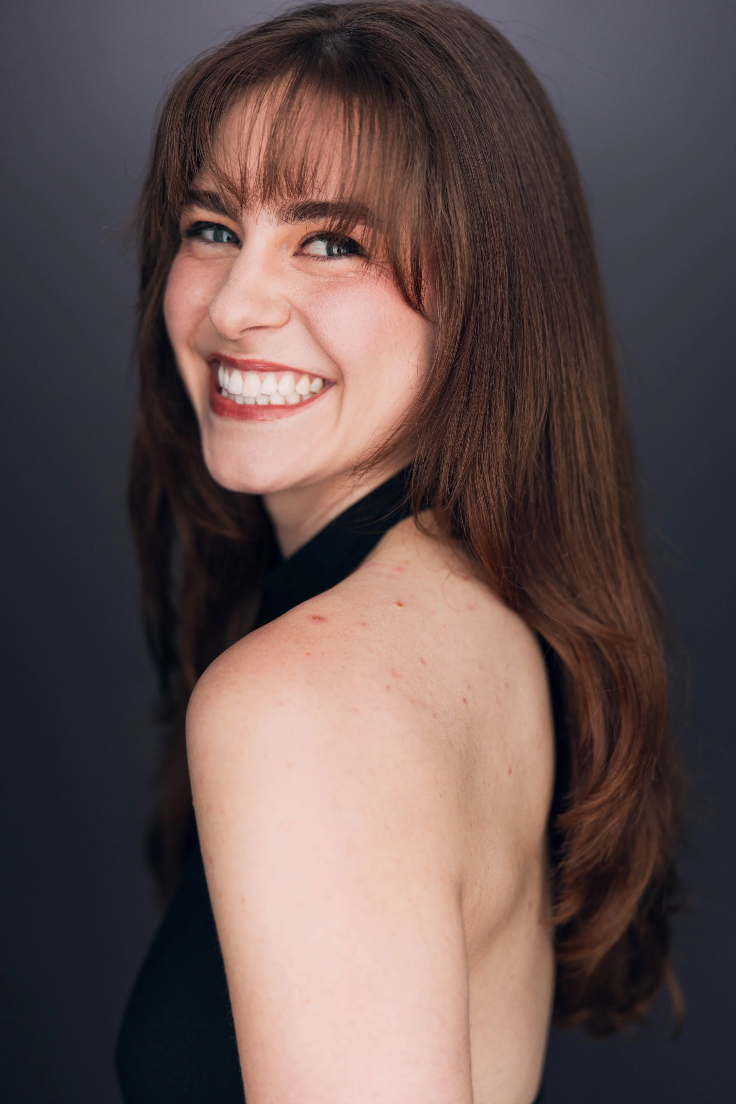 Close-up of a smiling woman with long, wavy brown hair, looking over her shoulder against a dark gray background.