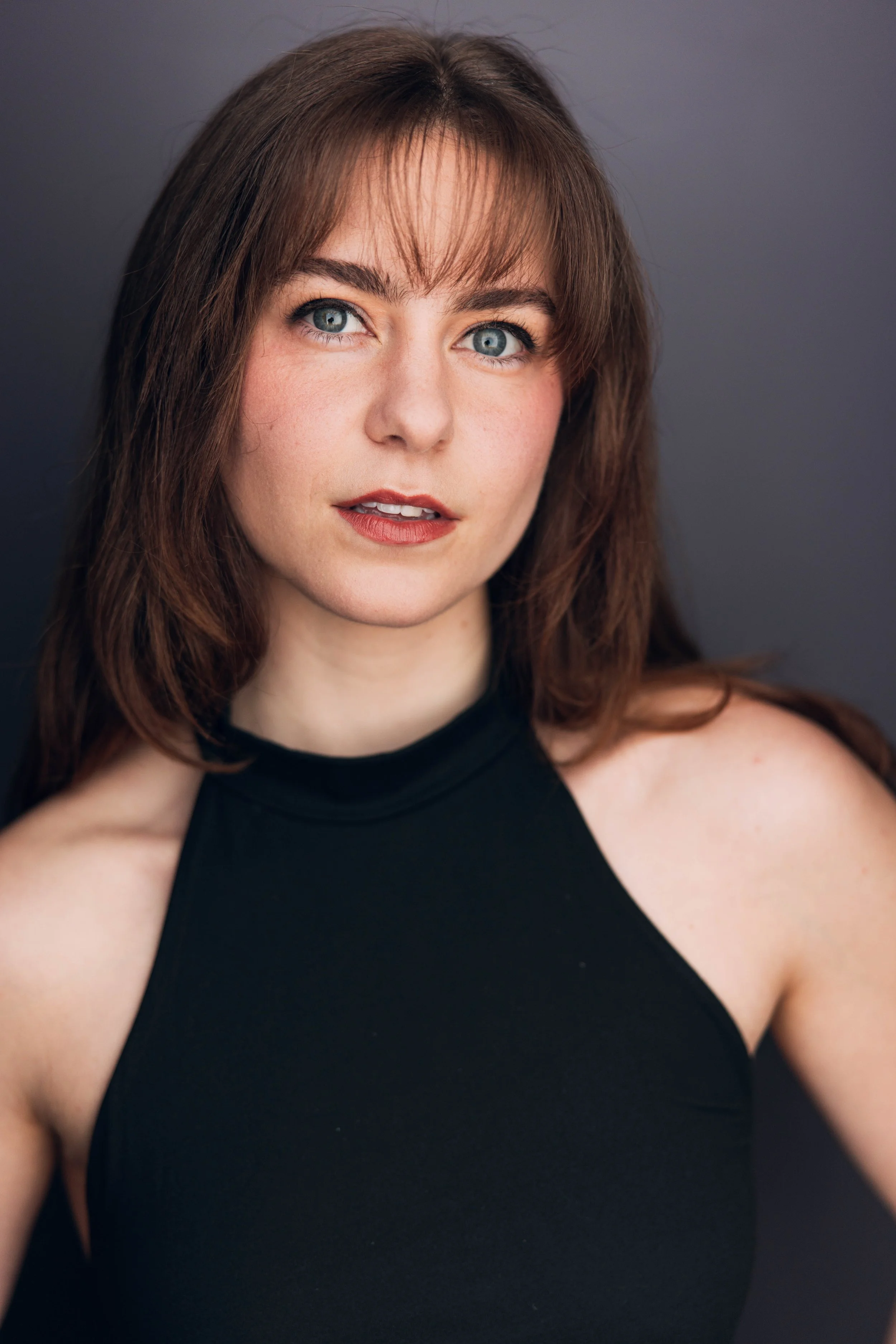 Close-up portrait of a woman with blue eyes and brown hair, wearing a black sleeveless top, against a gray background.