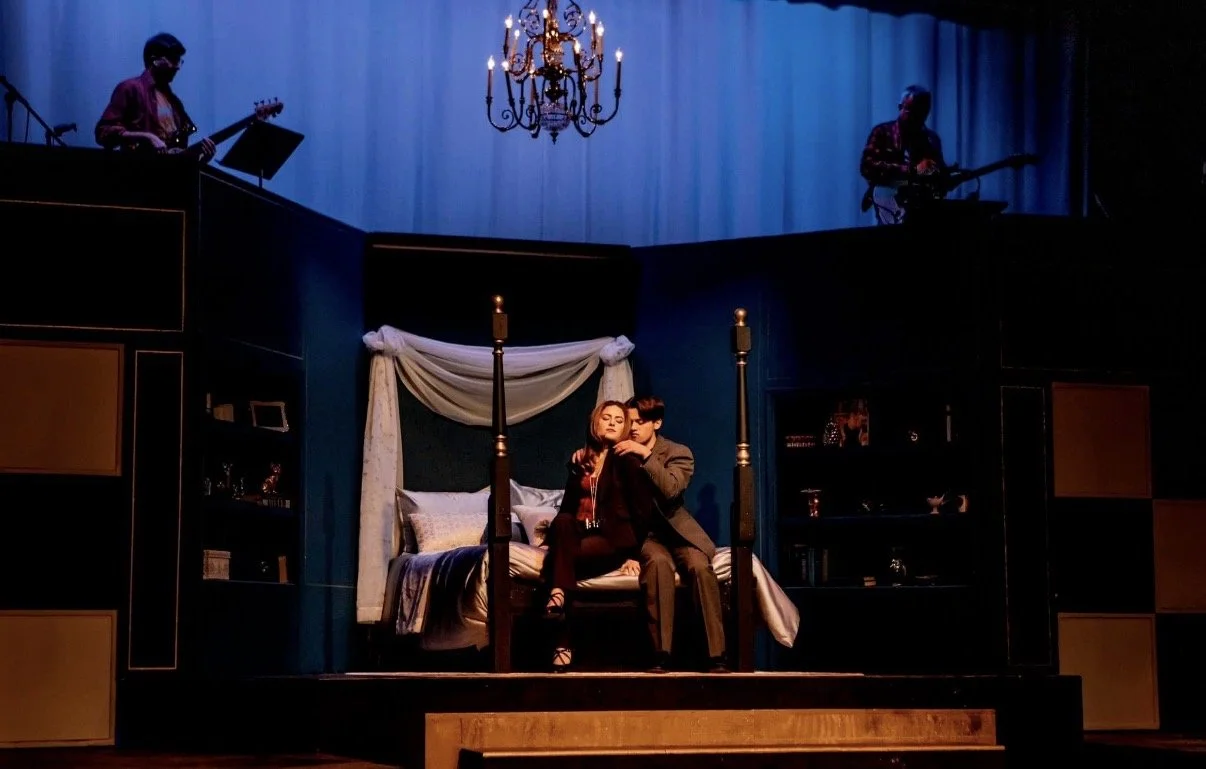 The scene shows a theatrical stage with a bedroom set, featuring a bed, two actors sitting on the bed, a hanging chandelier, and two musicians playing guitars in the background.