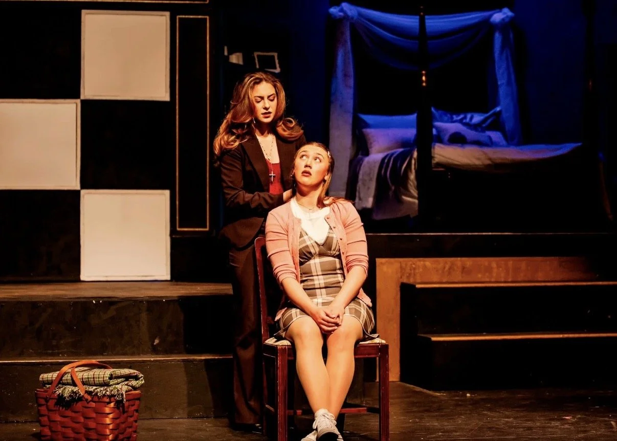 Two women on stage in a theatrical play, one sitting in a chair with a plaid dress and pink cardigan, the other standing behind her with a black blazer, in a room set with a bed and shelves.