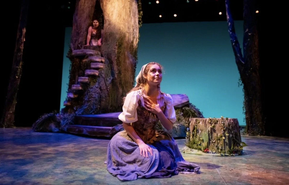 Two girls performing in a theater with a scene resembling a forest, one girl is kneeling with her hand on her chest, and the other girl is in a tree.