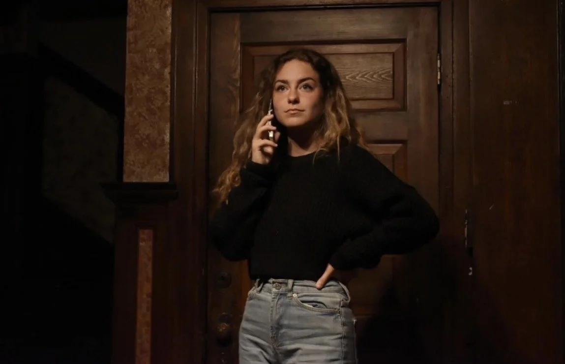 Young woman with curly hair wearing a black sweater and jeans, talking on a cellphone in front of wooden door.