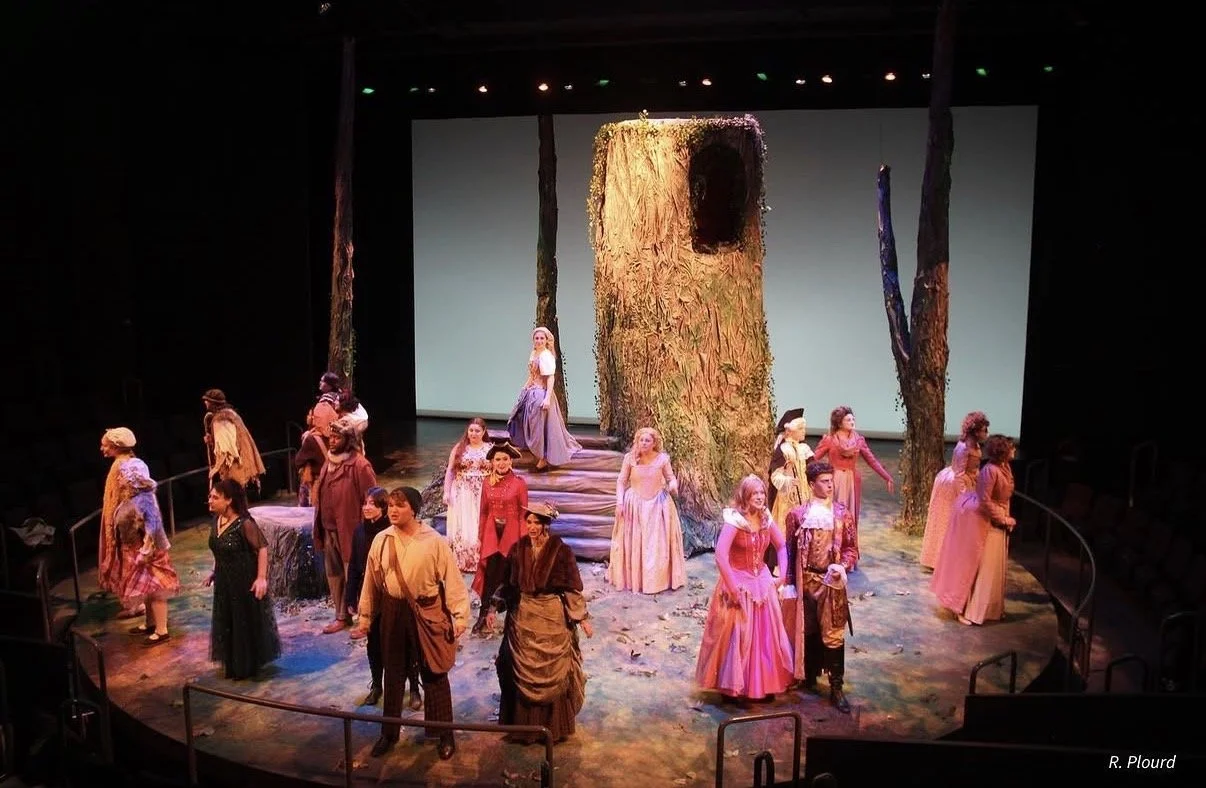 Actors on stage in a theater scene with a large tree stump and surrounding trees, dressed in period costumes, with a drone or large bird in the background.