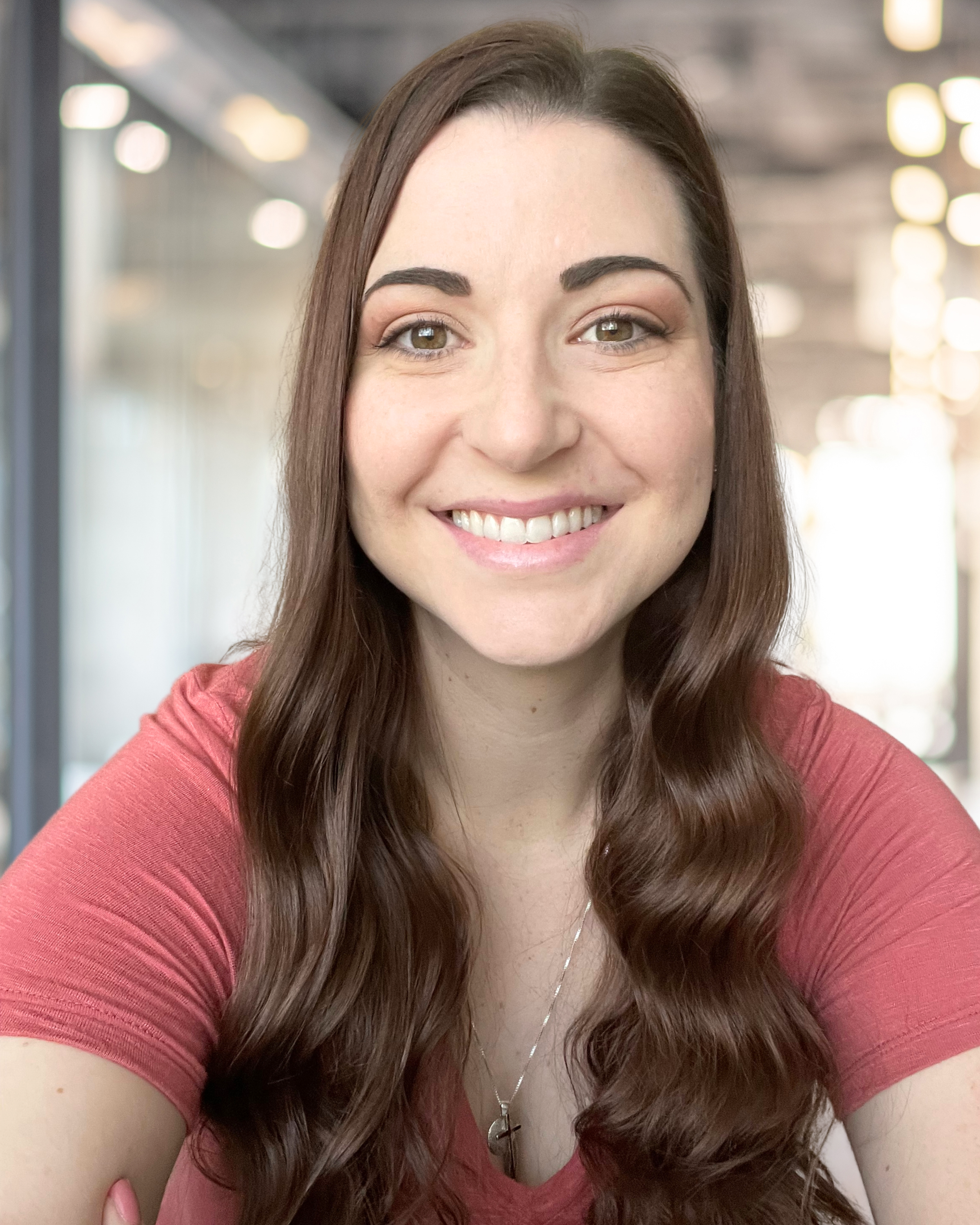 A woman with long brown hair, wearing a pink top and a silver necklace, smiling at the camera in an indoor setting with blurred background lights.