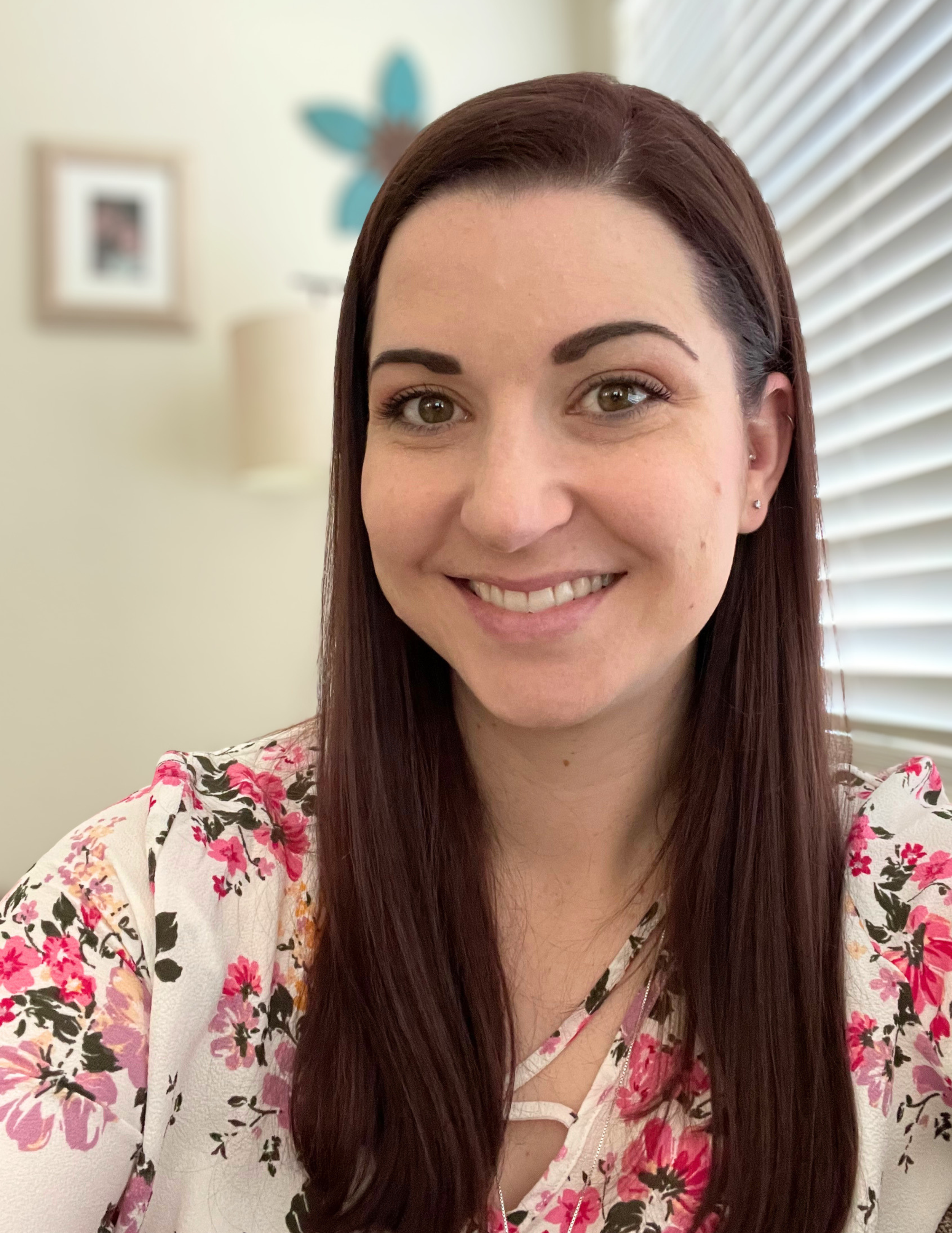 A woman with long, dark red hair smiling indoors near a window with blinds, wearing a white floral top.