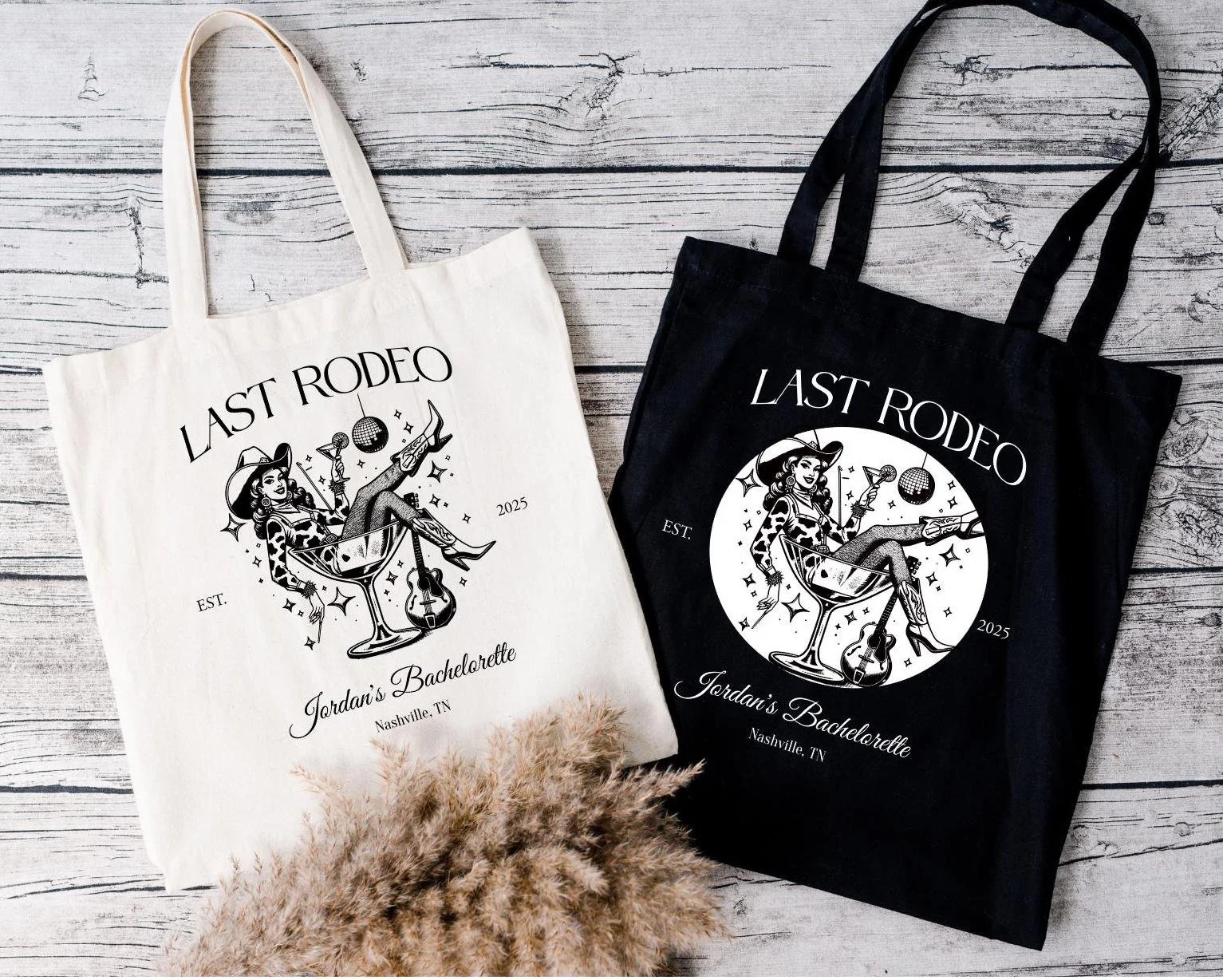 Two tote bags, one white and one black, with matching graphic designs of a woman sitting in a cocktail glass, holding a cocktail and a guitar, surrounded by stars, on a light wooden surface with dried pampas grass in the foreground.