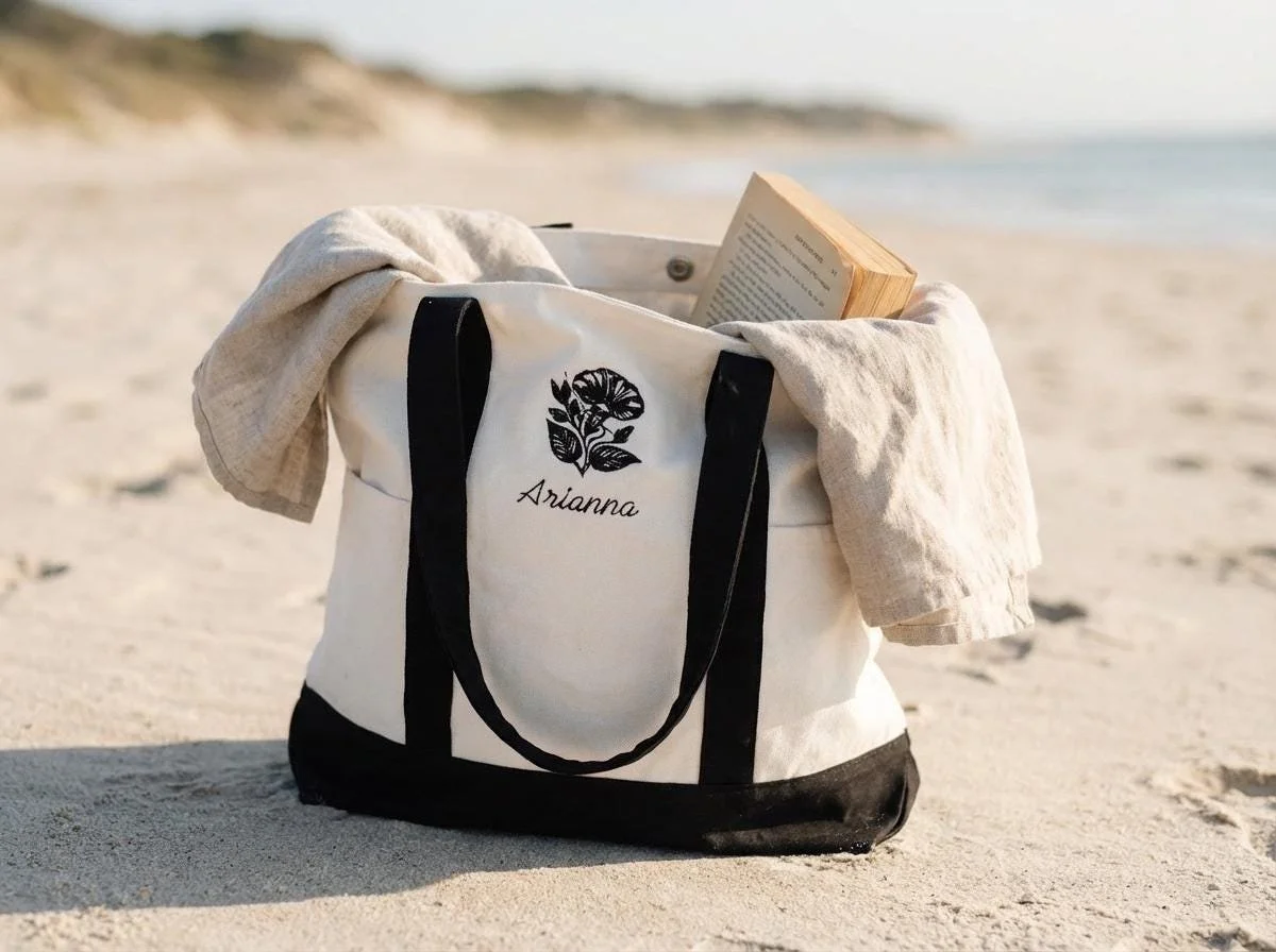 A white and black tote bag with the name 'Arianna' and a floral design on the beach, with a book and beige cloth inside and draped over the bag, sand and shoreline in the background.