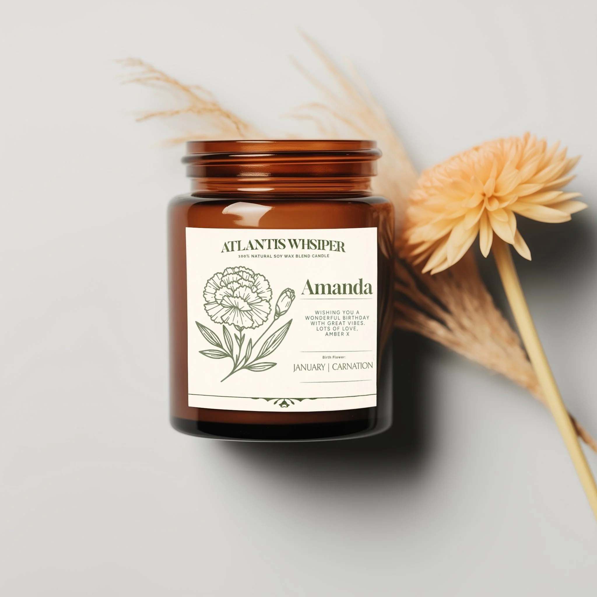 A brown candle jar with a white label that reads 'Atlantis Whisper'. The label features a line drawing of a flower and some text including 'Amanda' and mentions it is a natural soy wax blend candle. There is a beige flower and dried pampas grass beside the candle on a white surface.