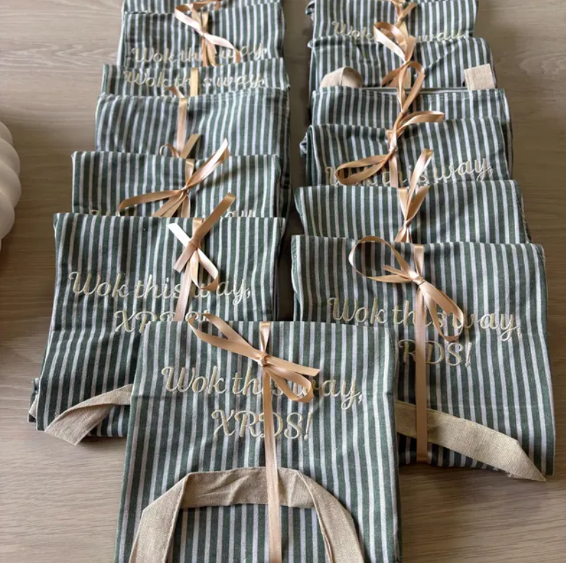 Multiple gift bags with striped green and white pattern, beige handles, and gold ribbons, arranged on a wooden surface. The bags have gold embroidered text reading "Wok this way, XPR'S!"